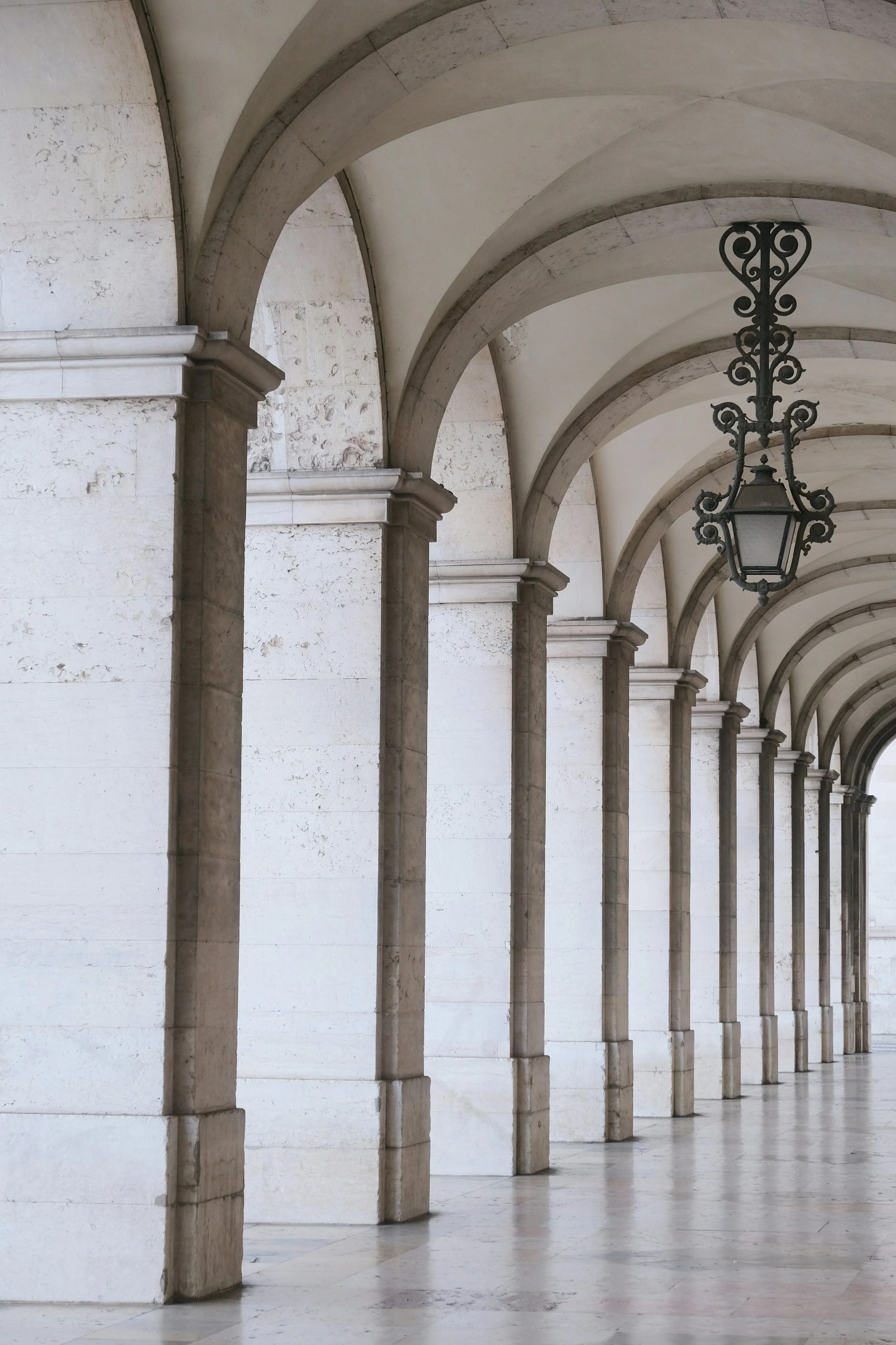 Daylight falls through a sequence of pale stone arches in a quiet colonnade. Photograph by Alex Kolundzija.
