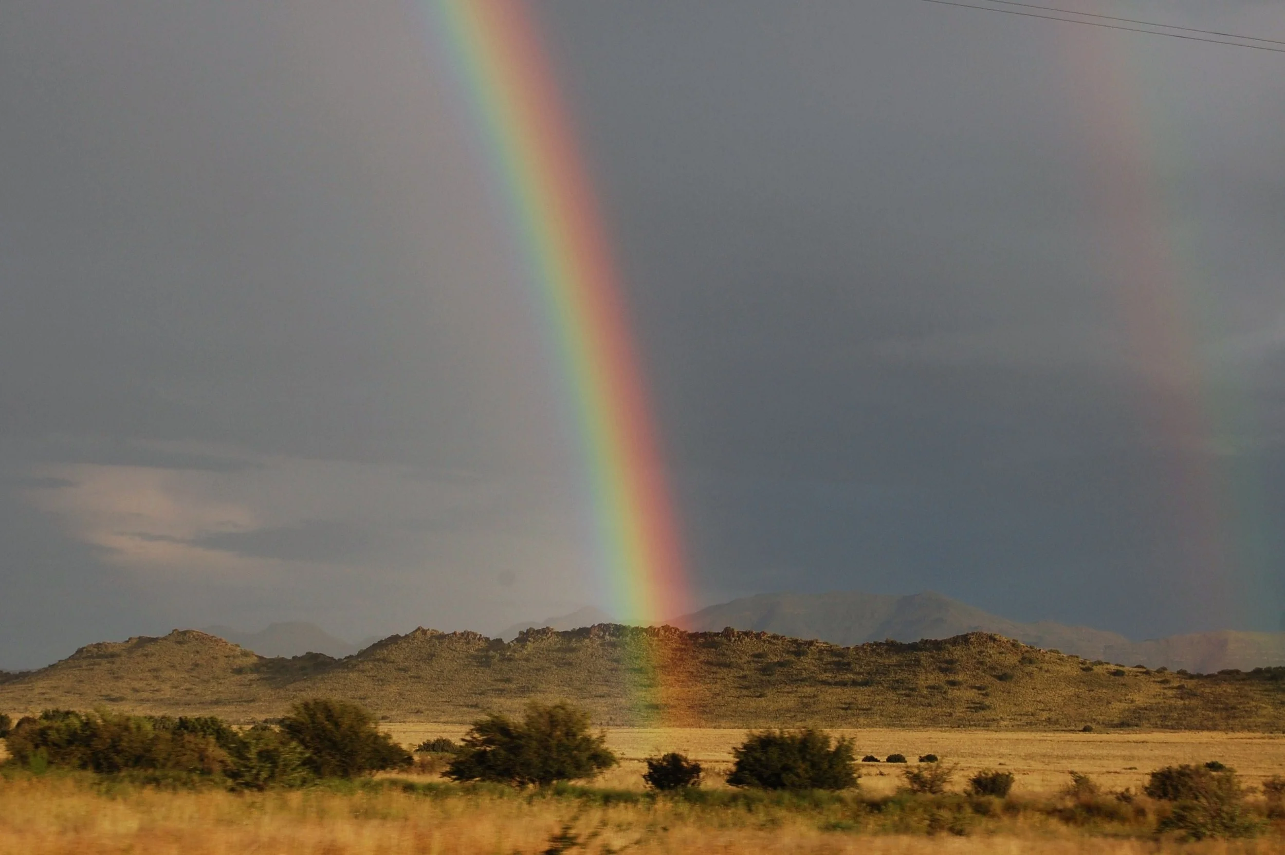 A rainbow arching across the sky over a hilly landscape with sparse vegetation and cloudy weather.