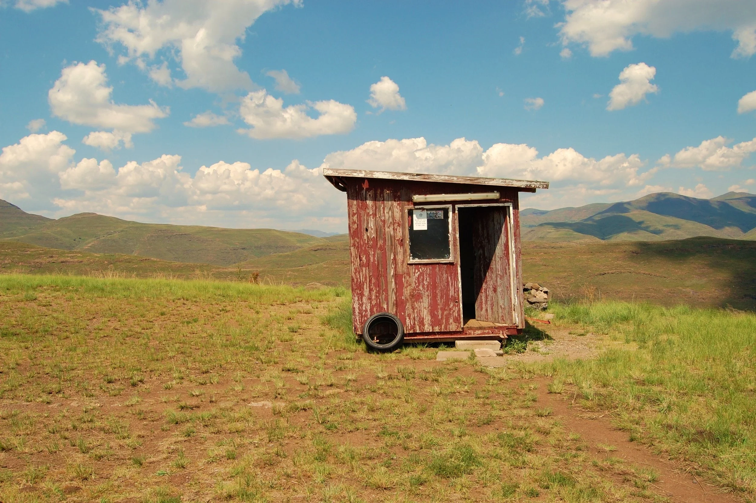 A small, weathered red shed with a slanted roof, standing alone in a grassy field with rolling hills in the background under a partly cloudy sky.