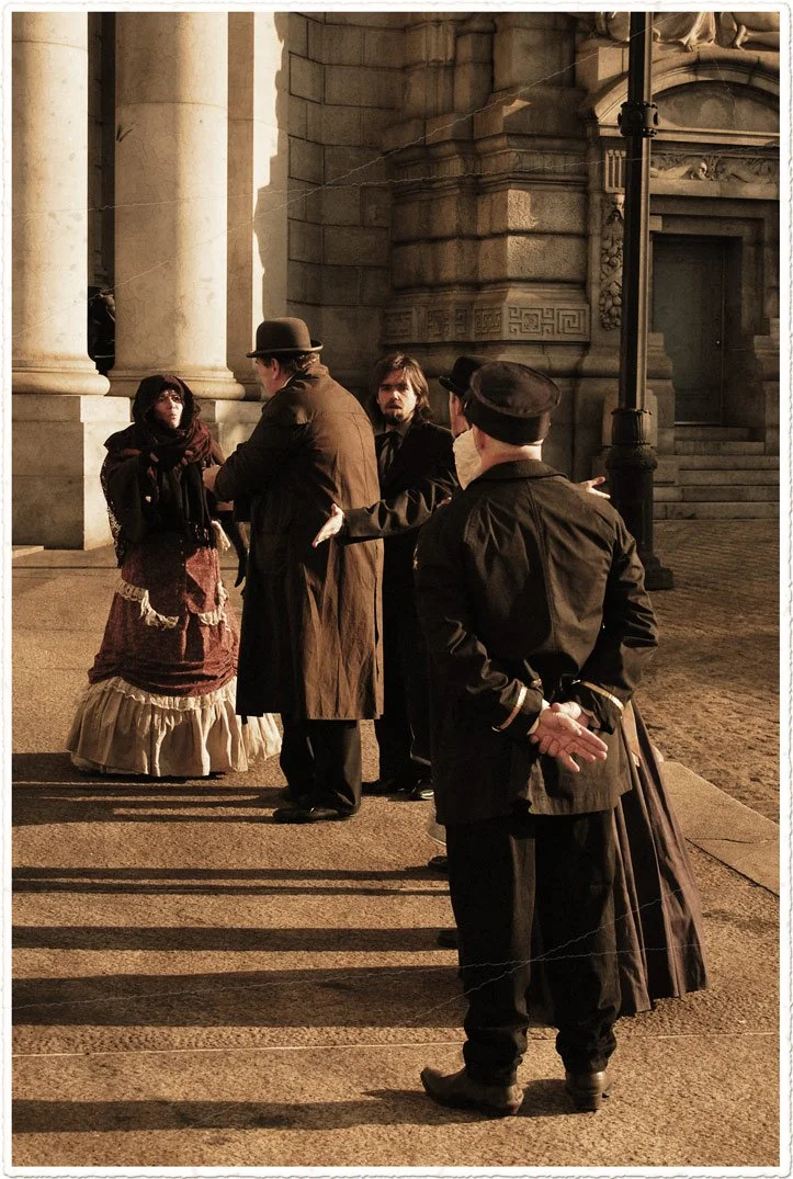 People dressed in vintage clothing, standing outside a historic building with large stone columns, possibly recreating a historical scene or participating in a film or themed event.