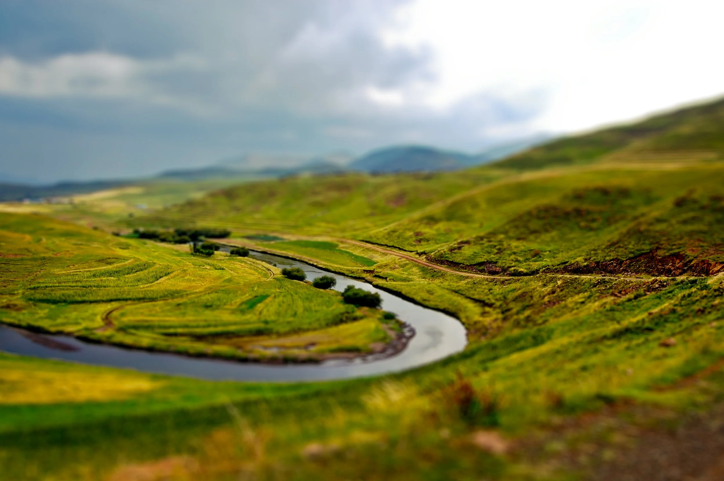 A winding river flows through green terraced fields in a hilly landscape with mountains in the background under a cloudy sky.