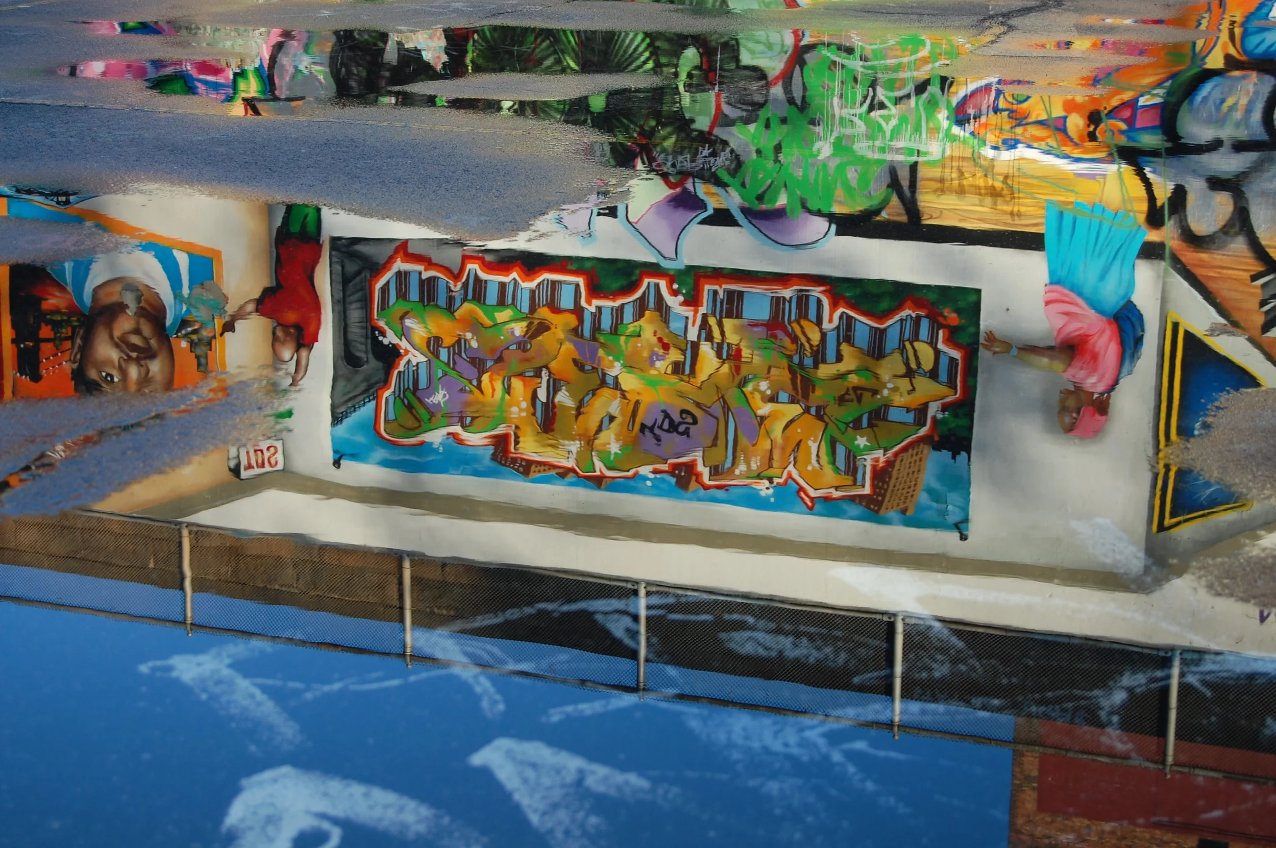 A colorful graffiti mural on a mural wall features vibrant street art with various designs and characters, including a face and abstract patterns, located outdoors with a blue sky in the background.