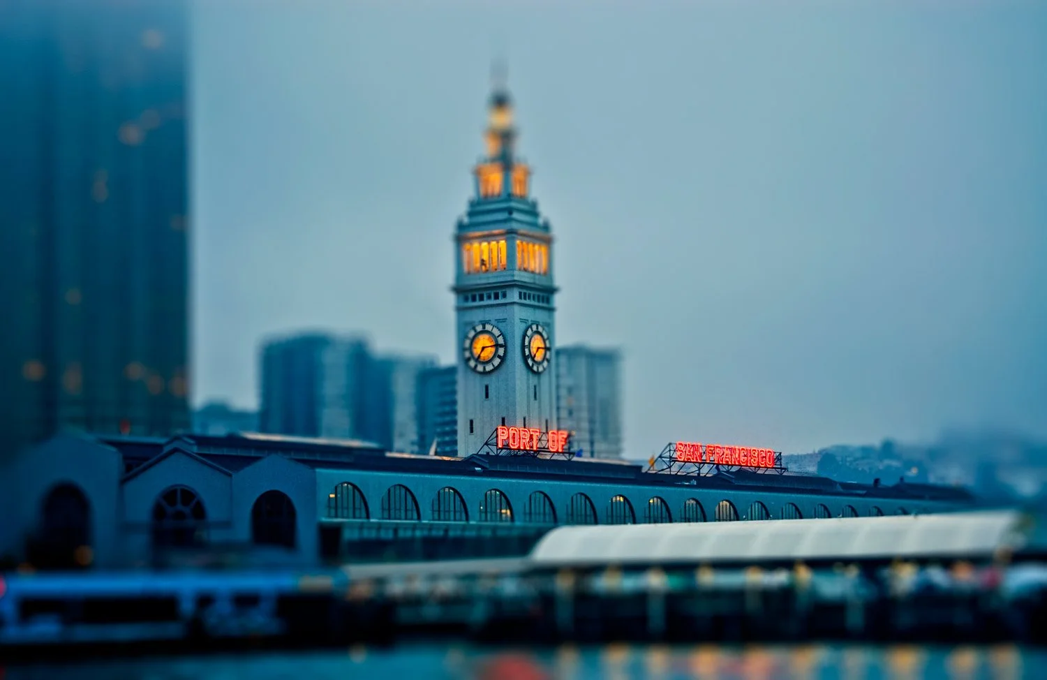 The clock tower at San Francisco's historic Ferry Building during dusk, with illuminated clock faces and a neon sign reading "PORT OF SAN FRANCISCO" in the foreground, and city buildings in the background.