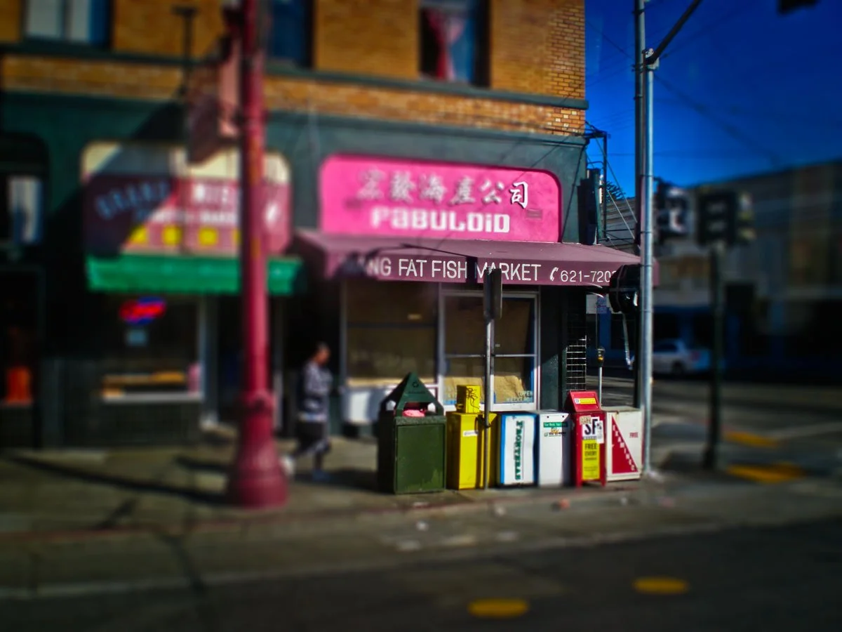 Street view showing a storefront with a pink sign, trash bins, and a person walking on the sidewalk.