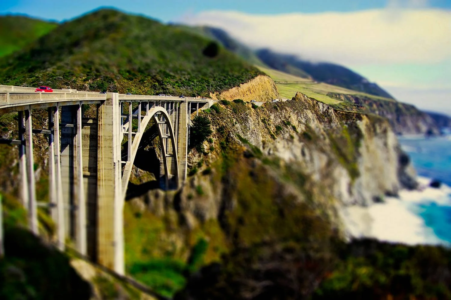 A coastal bridge with cars, set against green hills and rugged cliffs along the ocean.