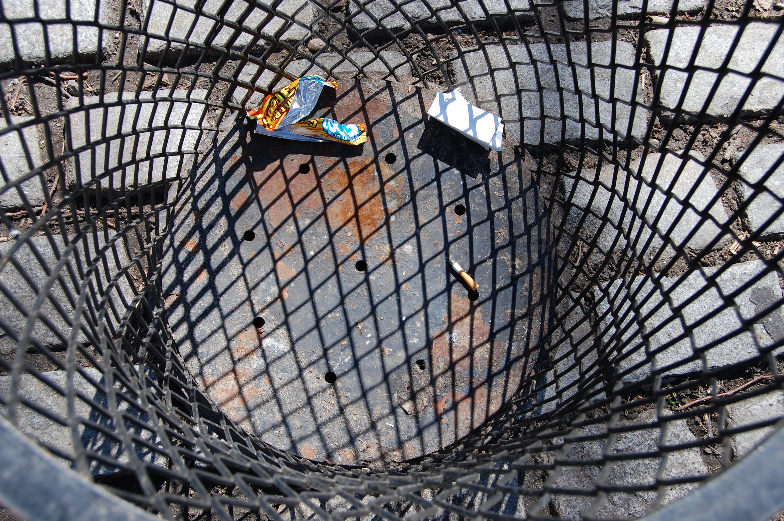 A black metal trash can with a diamond-patterned wire mesh, containing discarded items including a snack wrapper and a small white box, sitting on a concrete sidewalk with debris and dirt.
