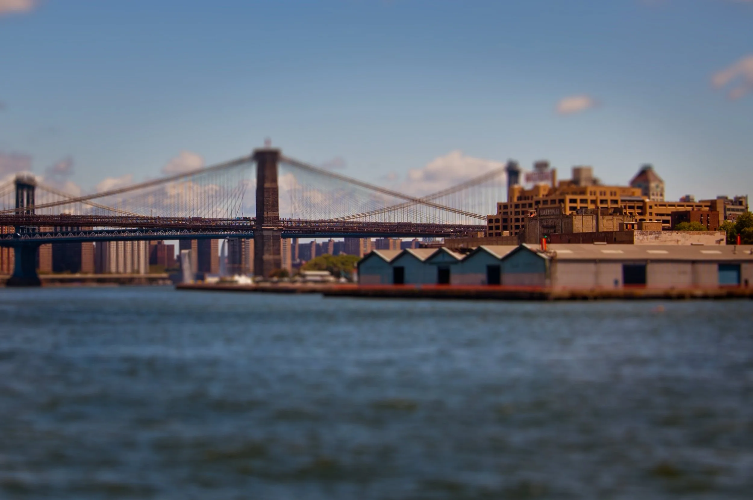 Blurred photograph of the Brooklyn Bridge with a city skyline in the background, taken from a vantage over the water.