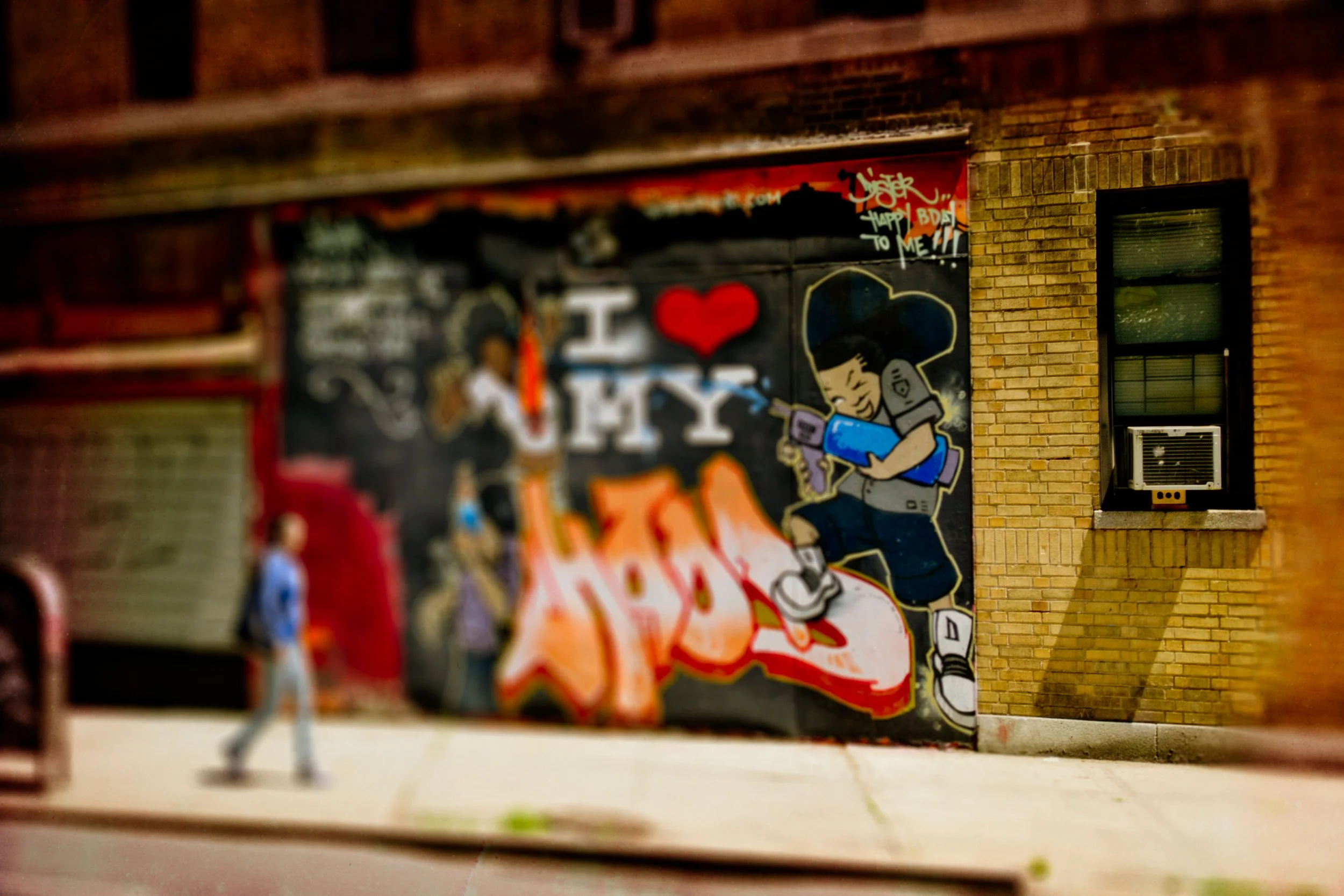 Street art mural with the text "I ❤️ NY" and a cartoon girl reading a book, located on a brick building wall.