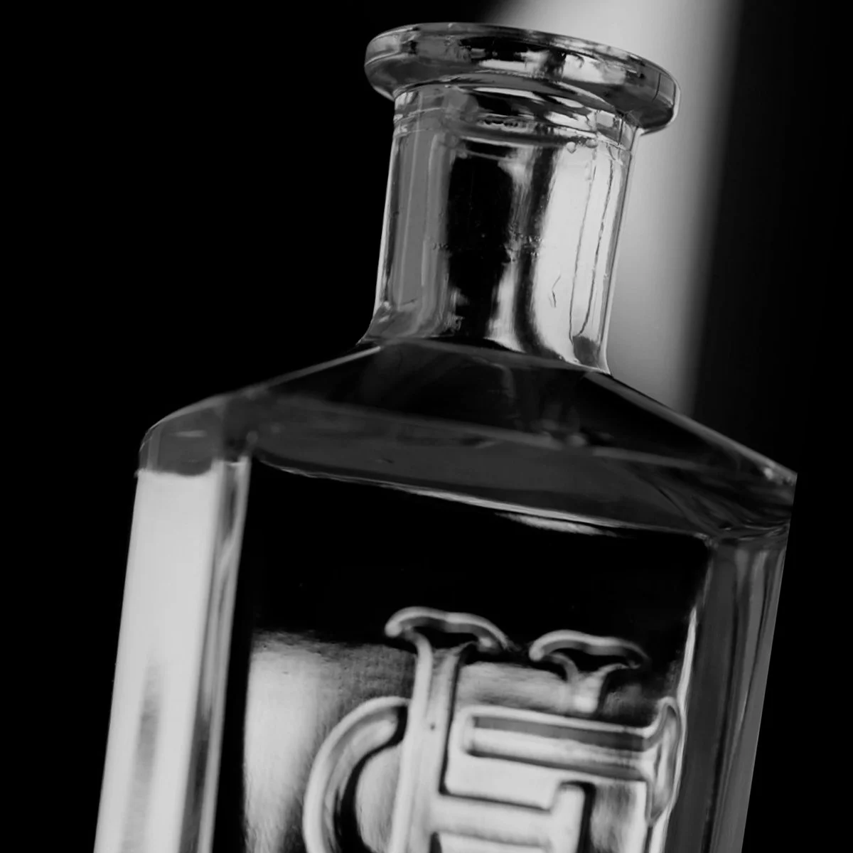 Close-up of a transparent glass bottle against a black background, showing the embossed brand logo on the front.