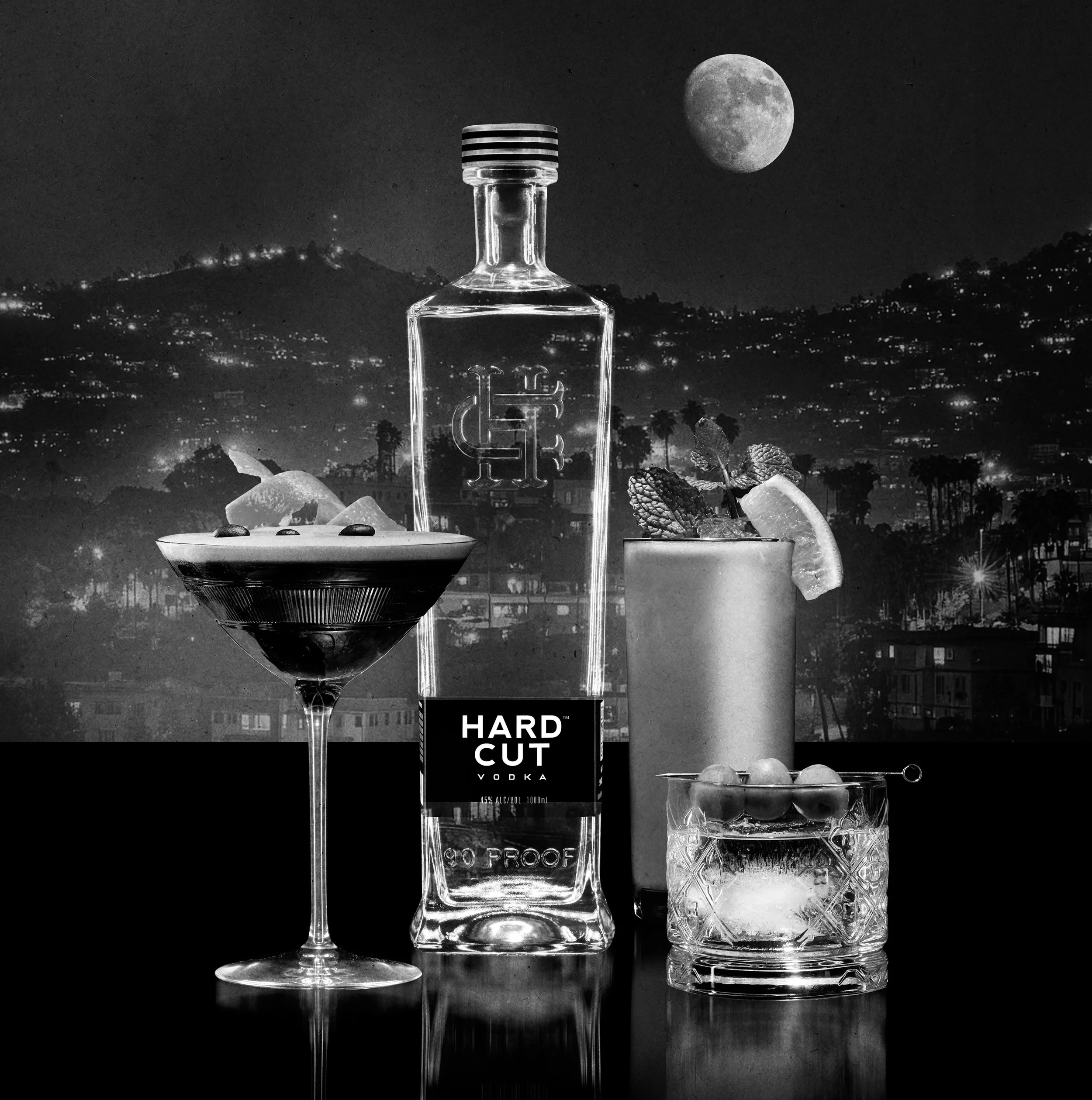 Black and white photo of a clear liquor bottle, a cocktail glass, and a glass of ice with mint leaves, with a full moon in the night sky in the background. The liquor bottle has dark caps, and the cocktail glass has an 'HARD GUT' label on it.