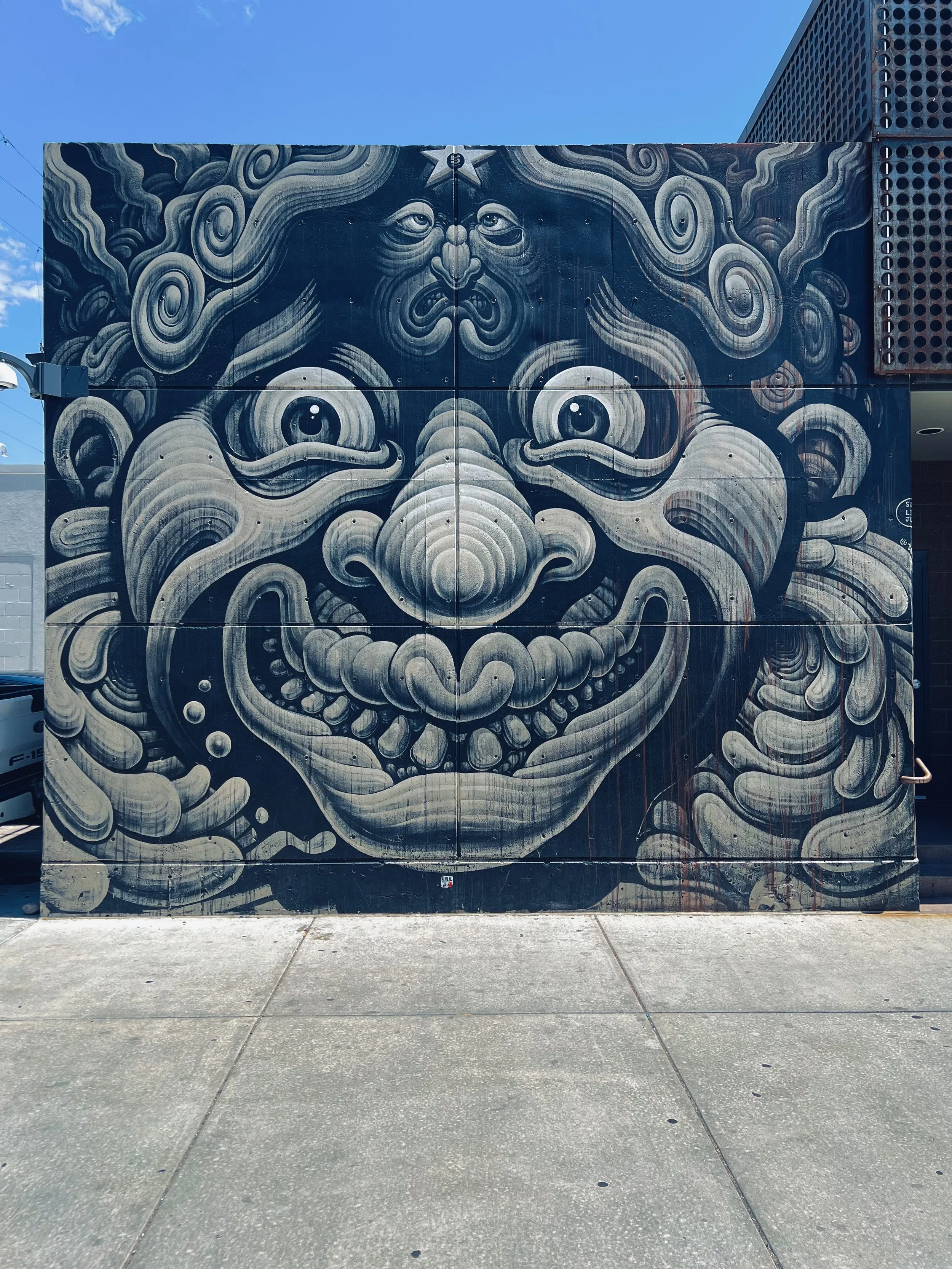 Street art mural of a stylized, fierce face with swirling patterns and a star on the forehead, painted on a building's exterior wall.