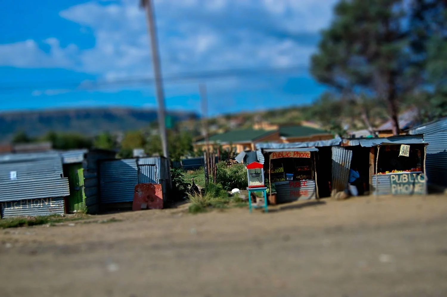 A dirt roadside with small makeshift shops made from corrugated metal sheets, including a public phone booth, under a blue sky with some clouds and distant trees.