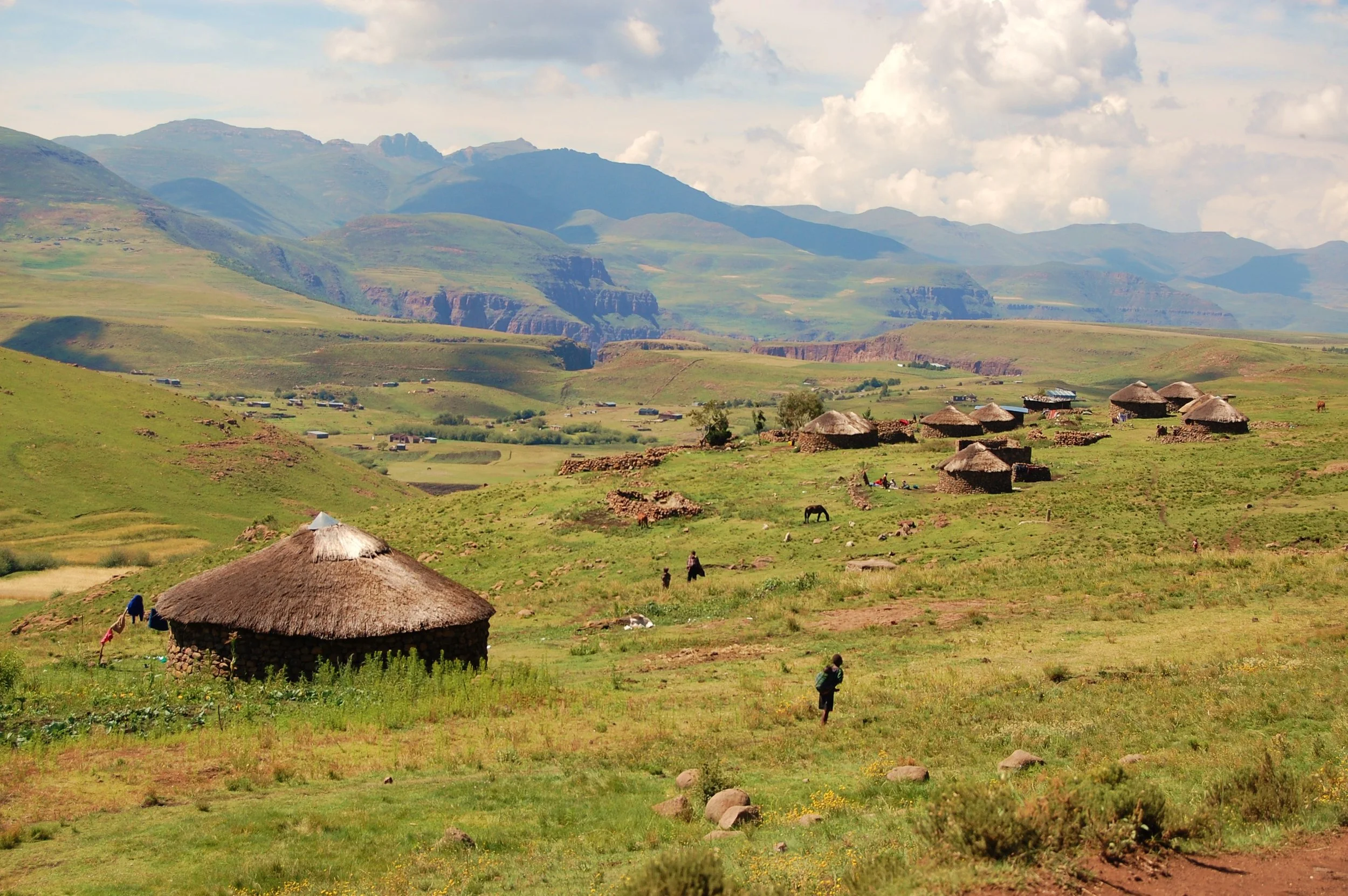A rural landscape with green rolling hills, traditional round huts with thatched roofs, scattered small buildings, and distant mountains under a partly cloudy sky.