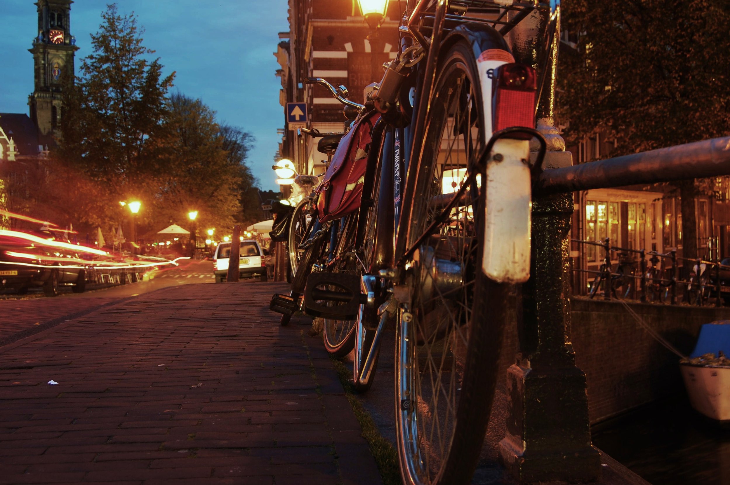Bicycles parked along a city street at dusk, with a church tower and illuminated street lamps in the background.
