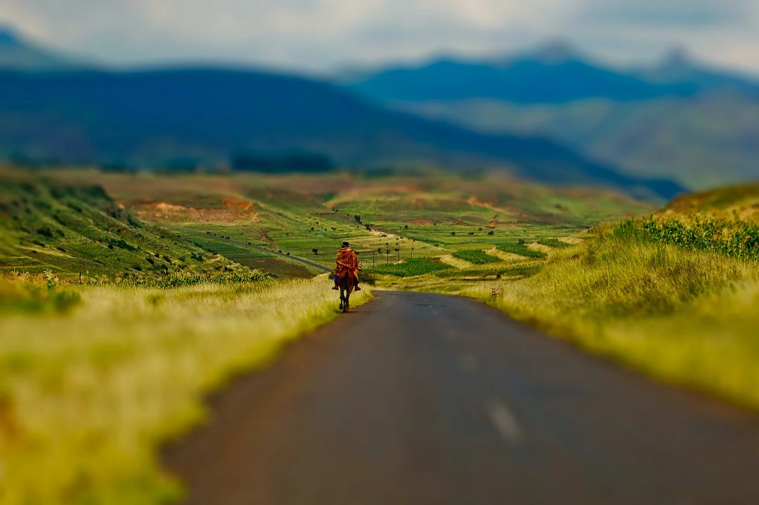 A person riding a horse along a rural road in a valley with green fields and mountains in the background.