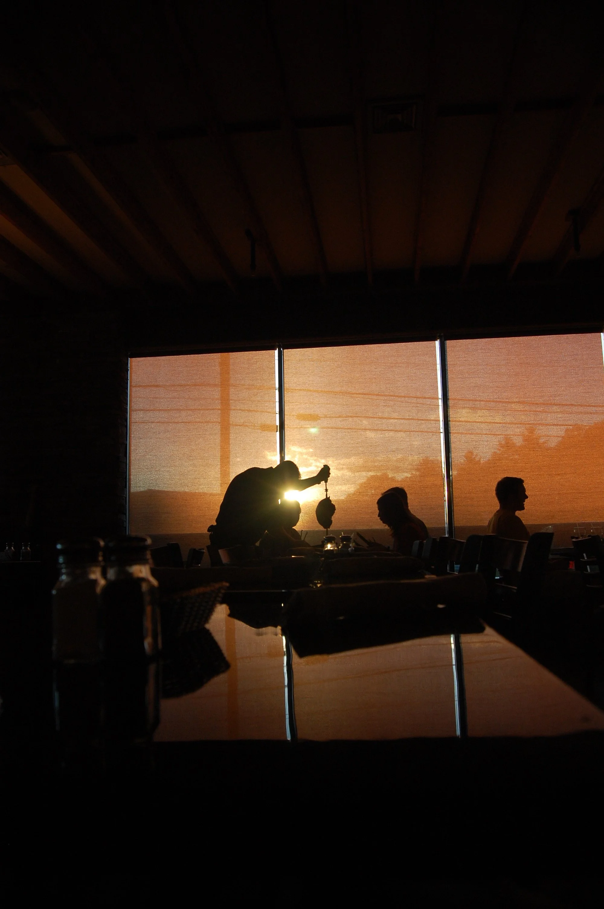 Silhouette of people dining at a restaurant during sunset with orange roller shades and wooden ceiling.