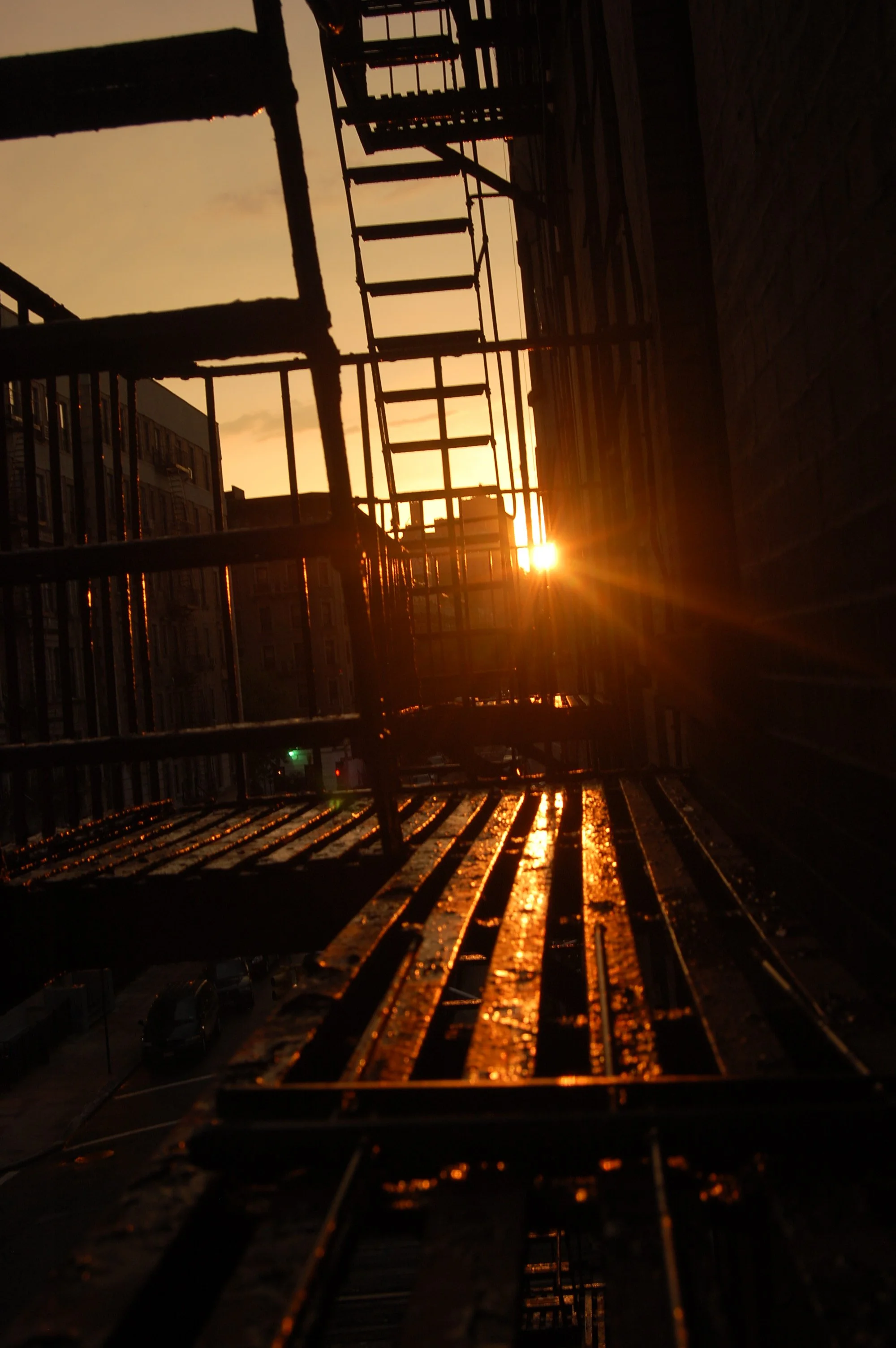 Sunset view through the metal fire escape stairs of a building, with warm orange lighting and silhouettes of surrounding buildings in the background.