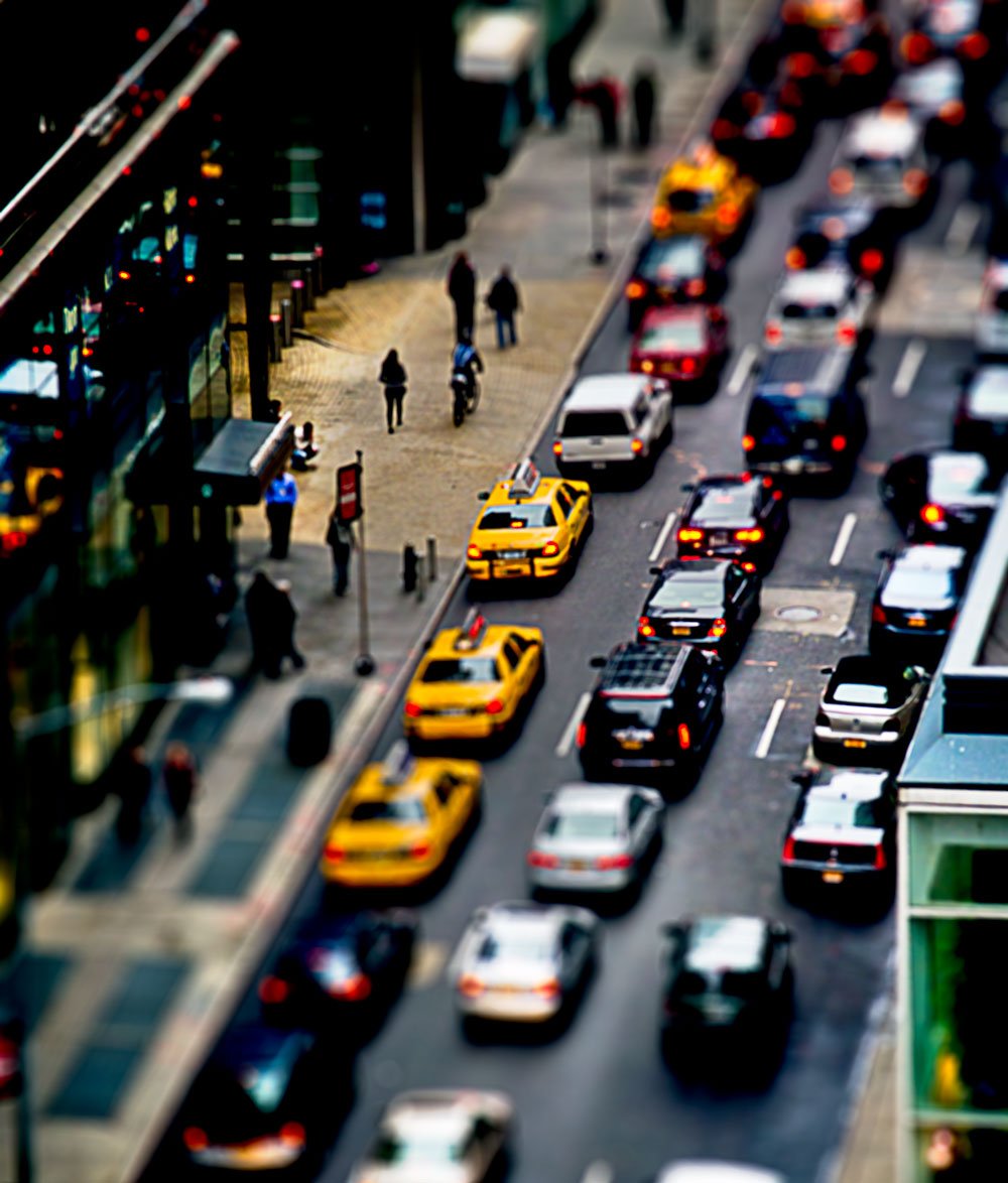 A bustling city street with traffic congestion, including yellow taxis, black cars, and white vehicles. Pedestrians walk along the sidewalk, and a storefront with bright lights is visible on the left side of the image.