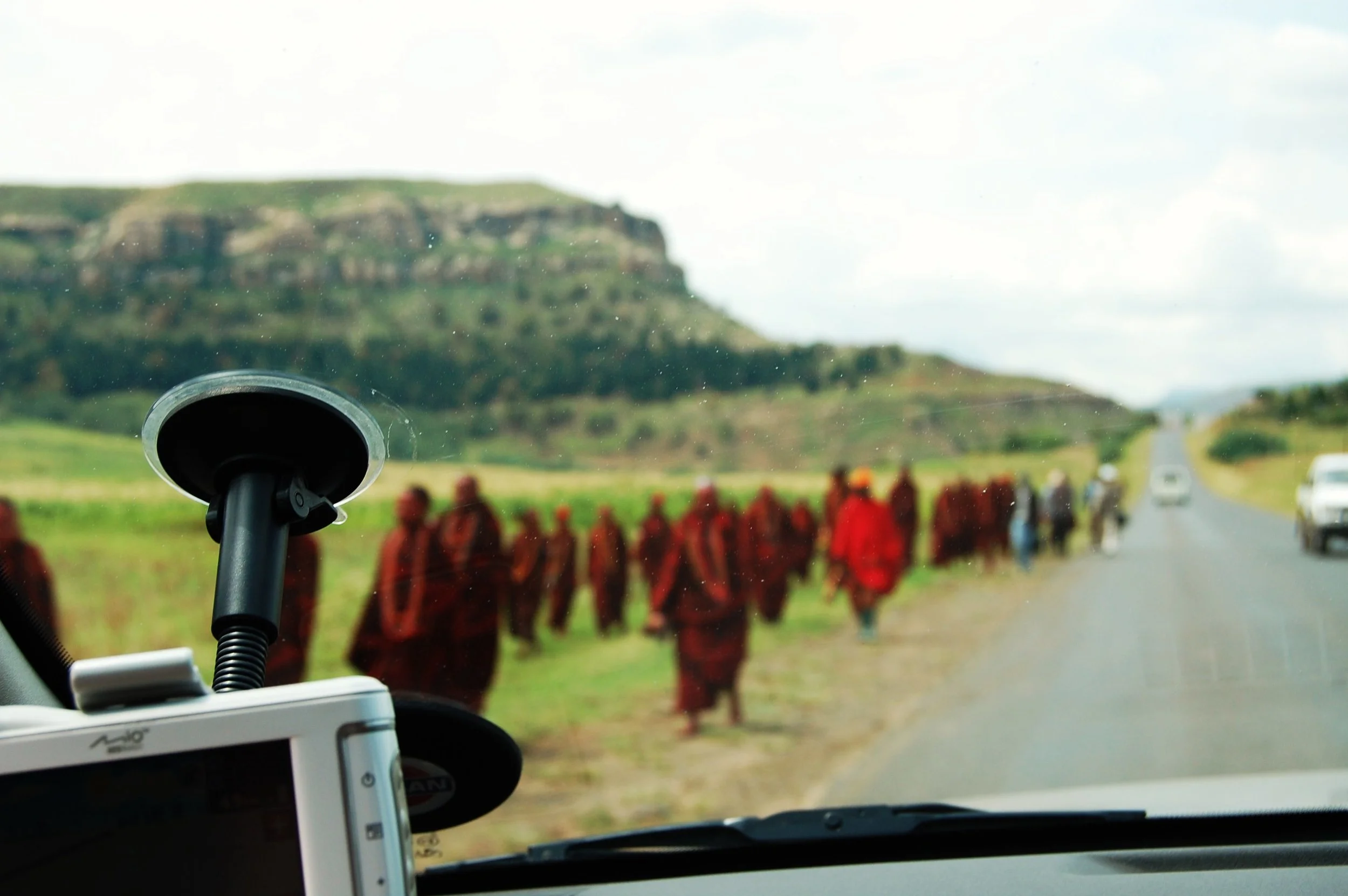 Photo taken from inside a vehicle showing the dashboard with a GPS and a camera, looking through the windshield at a group of monks walking along a rural road with green hills in the background.
