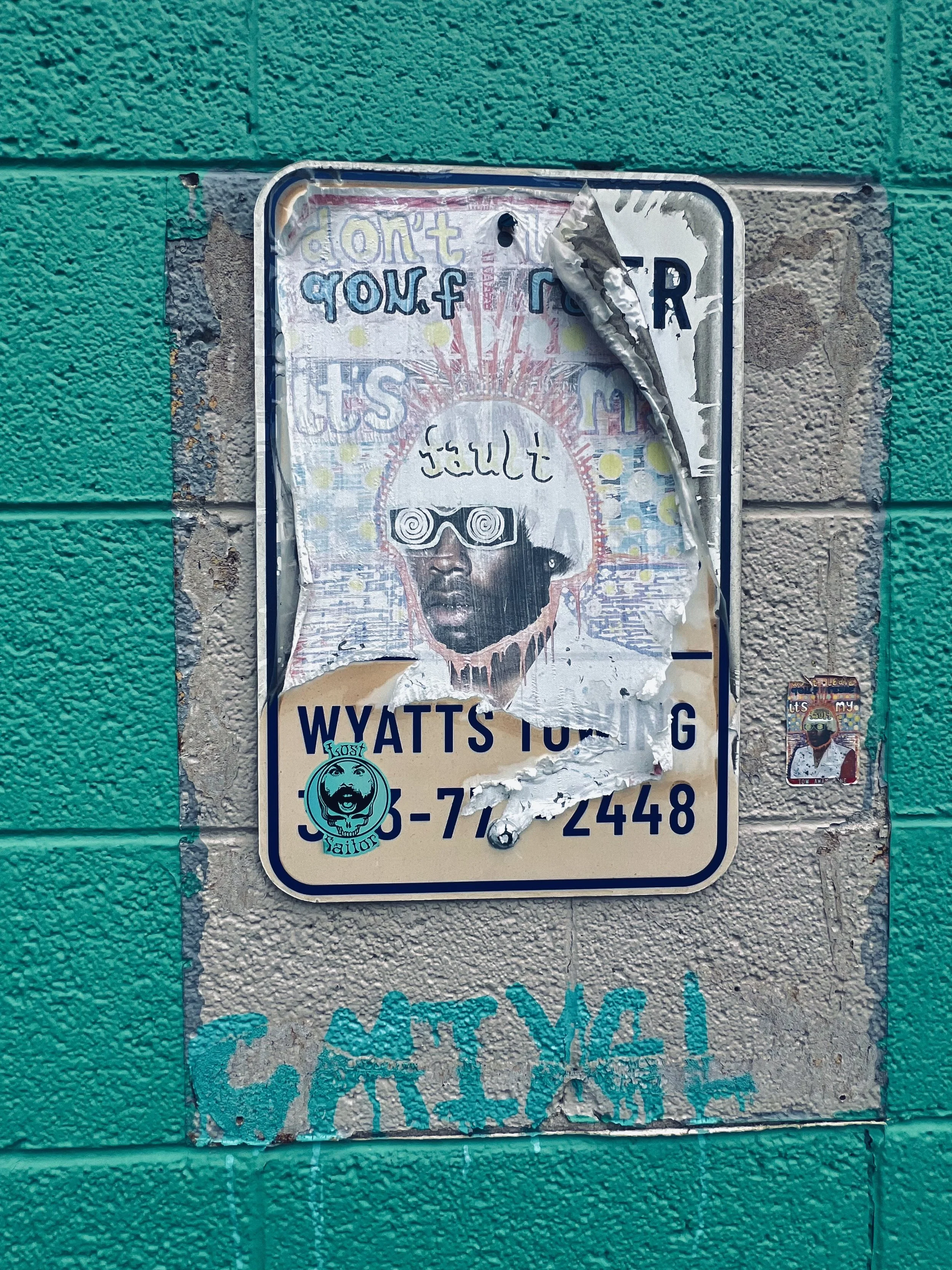 Torn and damaged parking or warning sign attached to a textured wall that is painted in turquoise and beige. The sign has text and images that are not fully legible, with additional smaller stickers or posters, including one featuring a person wearing sunglasses and a helmet.