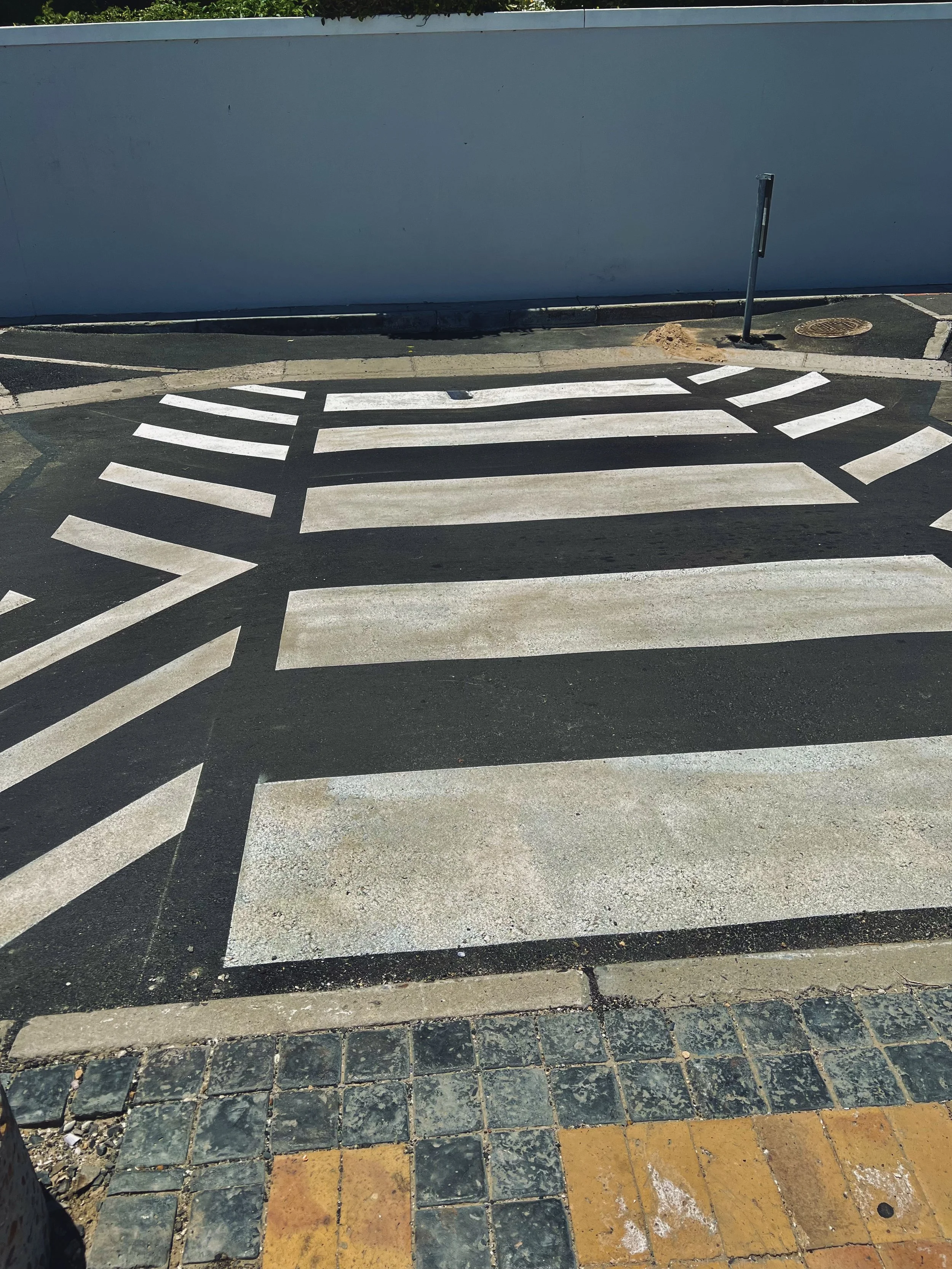 A pedestrian crosswalk with white stripes on asphalt, a sidewalk with black and gray cobblestones, and a curb in front. There is a blue-gray wall in the background, with a bent parking meter leaning to one side, and a small pile of dirt near its base. A circular metal utility cover is also visible on the street.