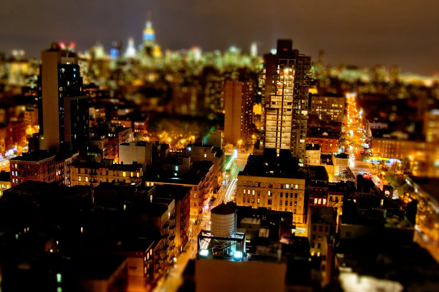 Nighttime cityscape with illuminated buildings and streets, some high-rise structures, blurred lights, and a recognizable building with a dome in the distant background.