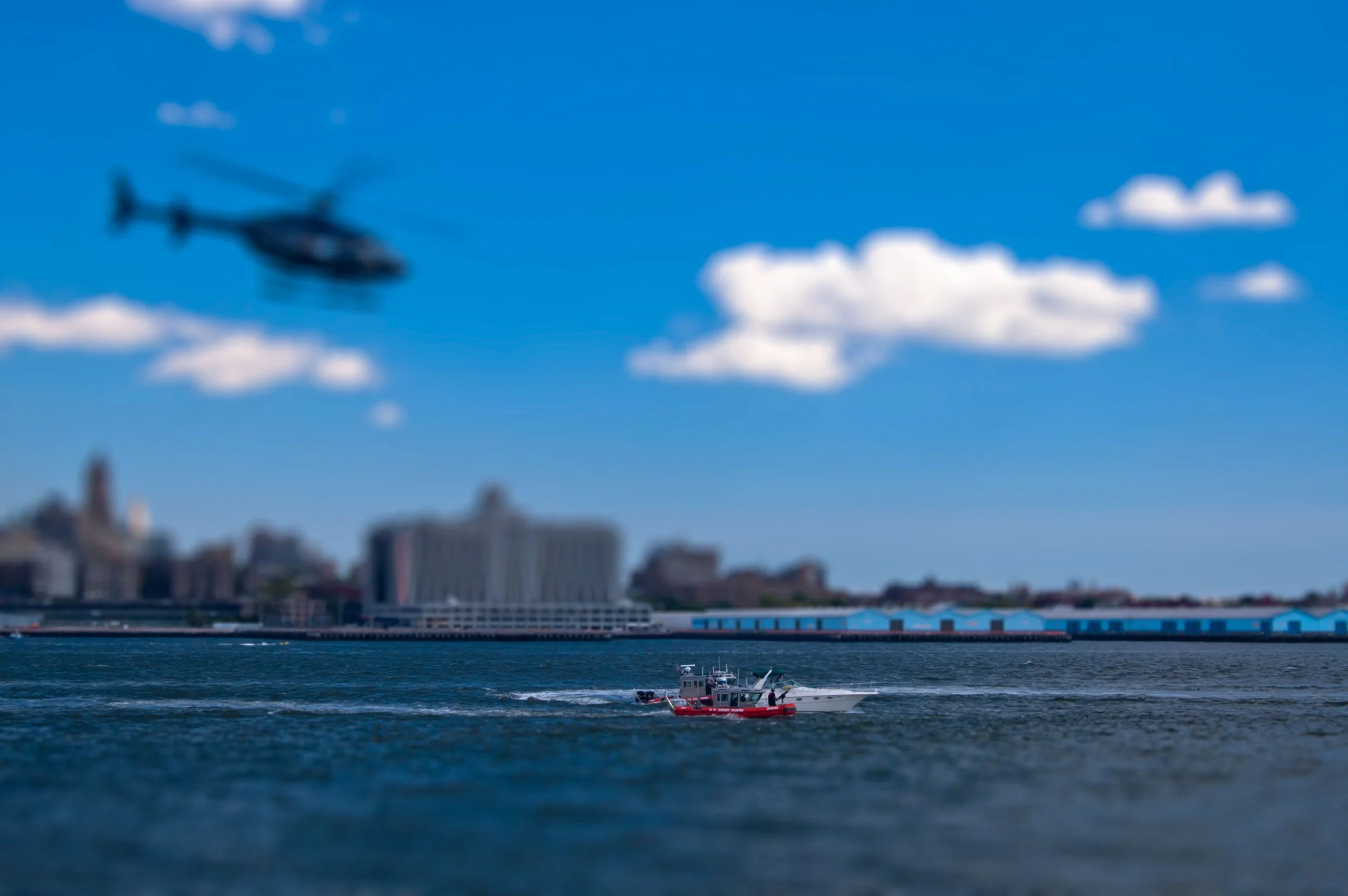 A blurry helicopter flying in the blue sky with some white clouds, above a cityscape near the water, with boats and buildings in view.