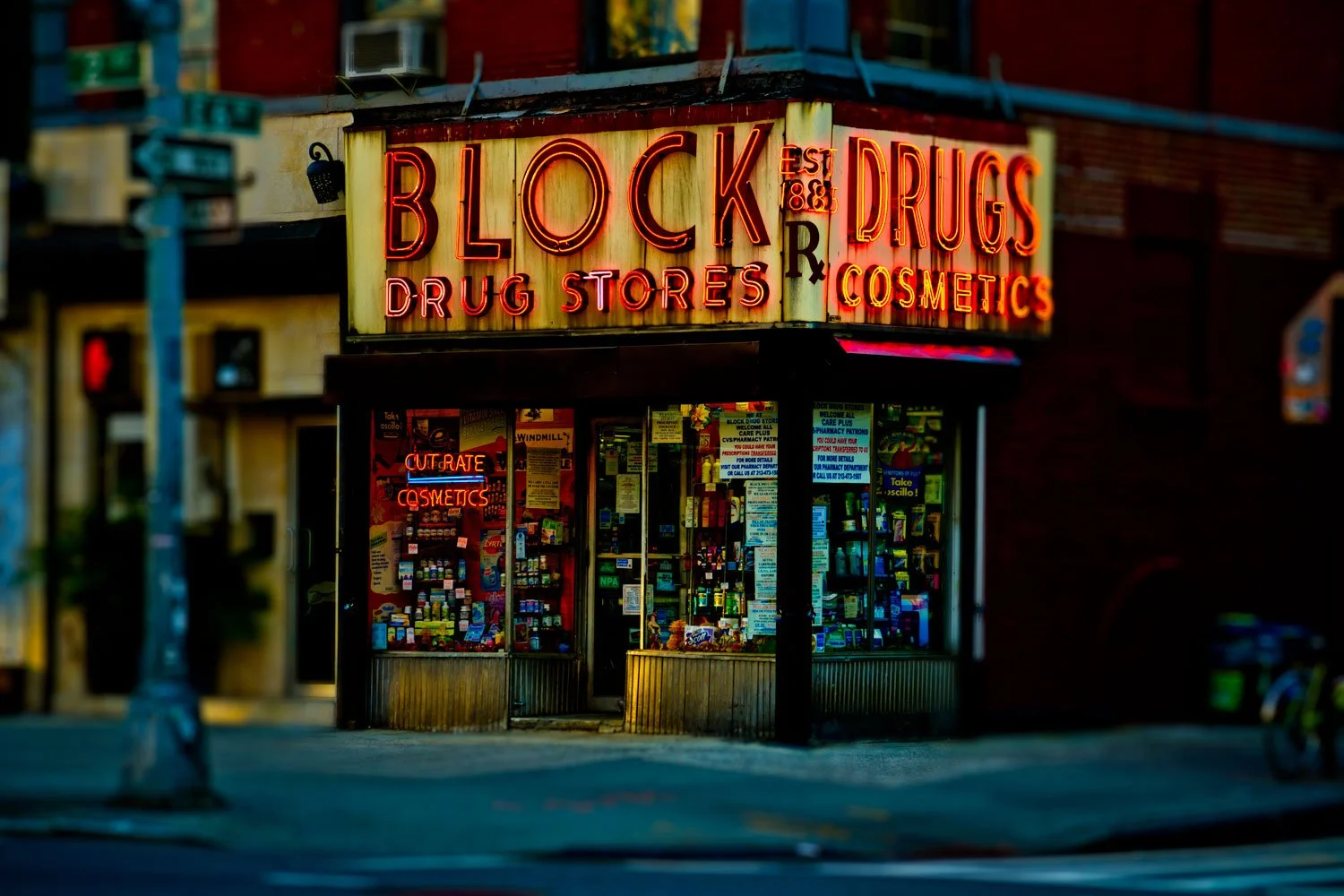Nighttime view of a drugstore with neon signs reading 'BLOCK DRUGS' and 'DRUG STORES', displaying various advertisements and products inside.