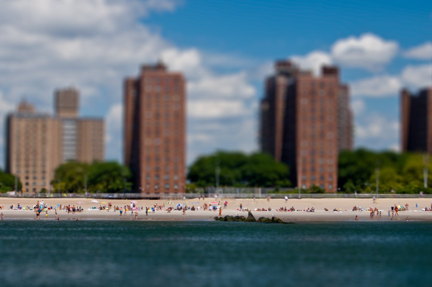 Beach with people sunbathing, swimming, and walking along the shoreline, with large apartment buildings and green trees in the background under a partly cloudy sky.