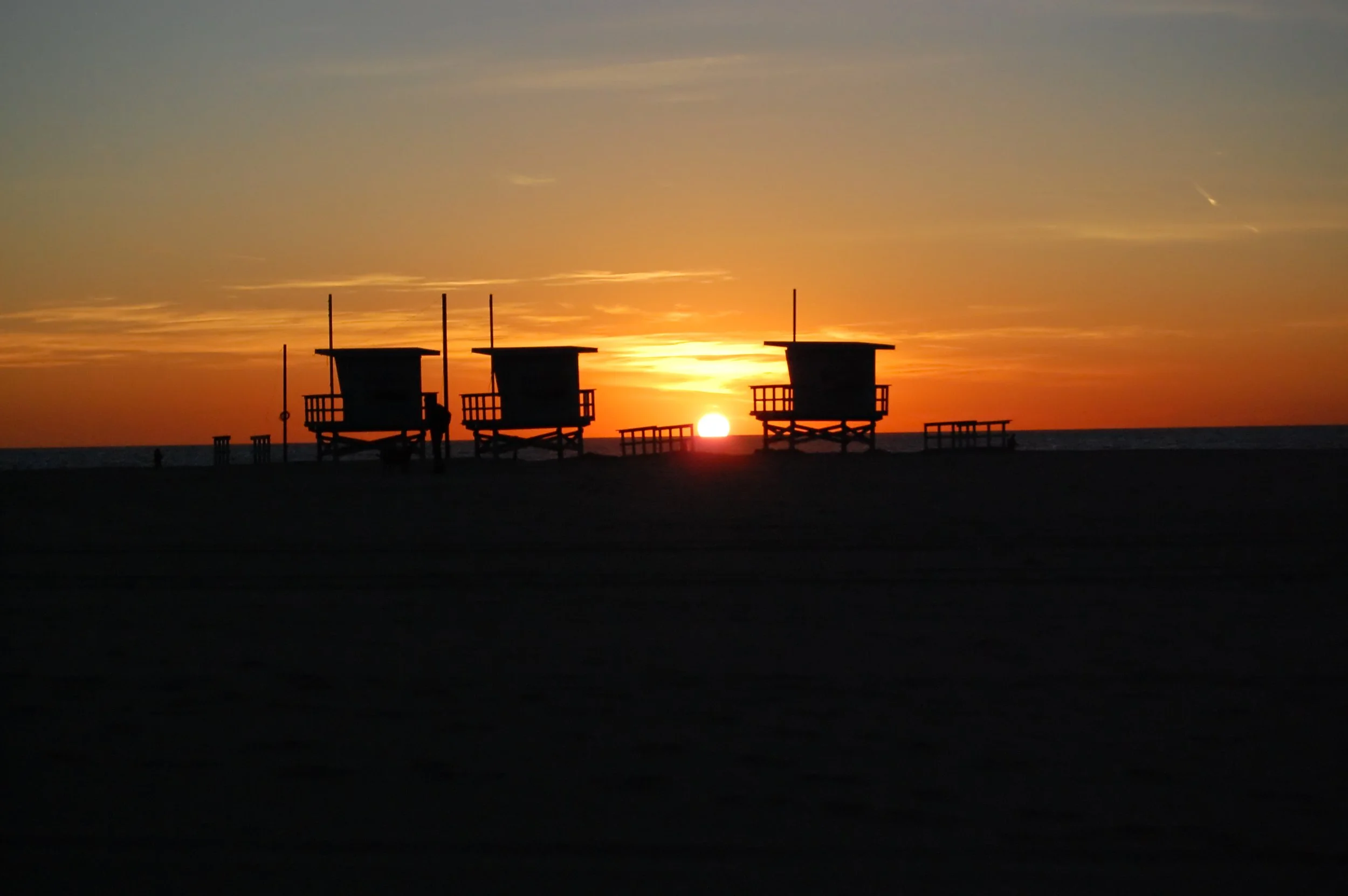 Silhouetted lifeguard towers along the beach at sunset with a vibrant orange and yellow sky.