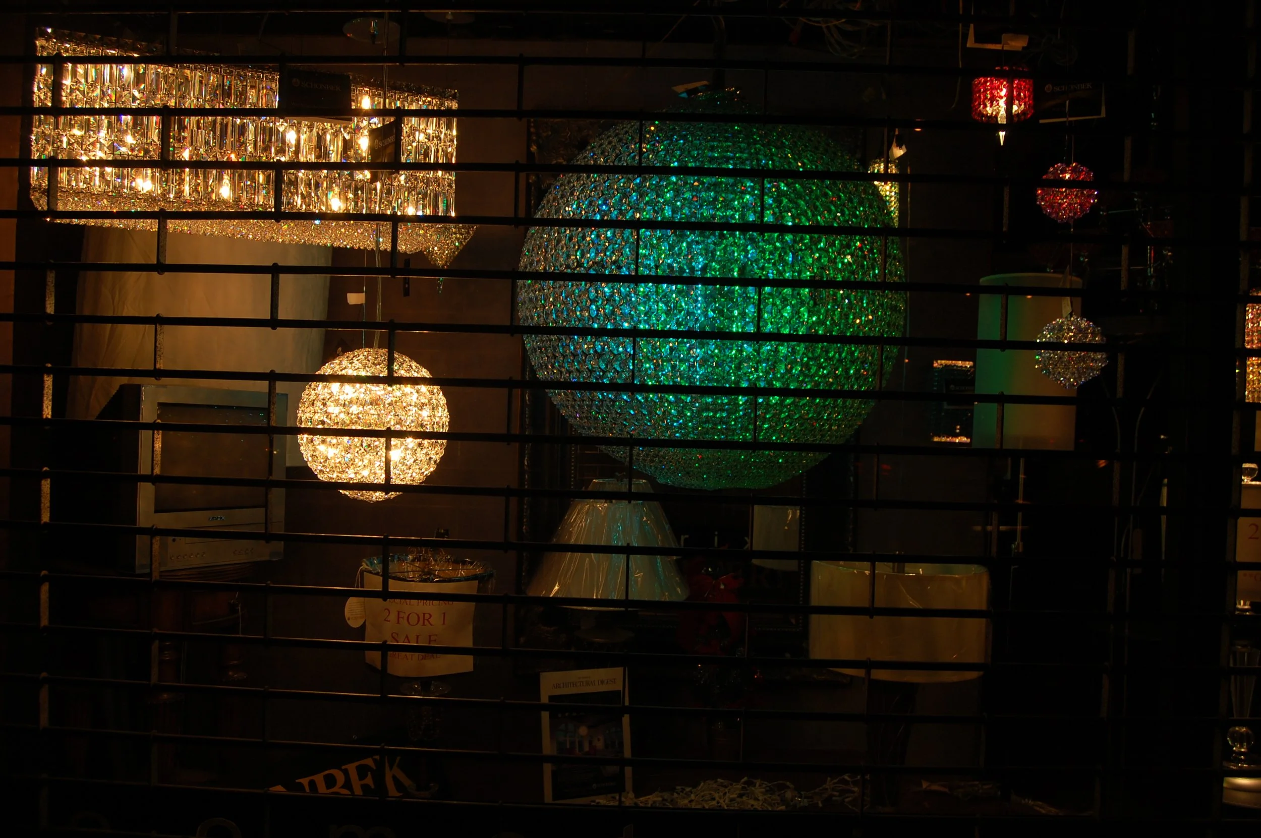 Various decorative chandeliers and hanging lamps displayed behind a window with black horizontal bars.