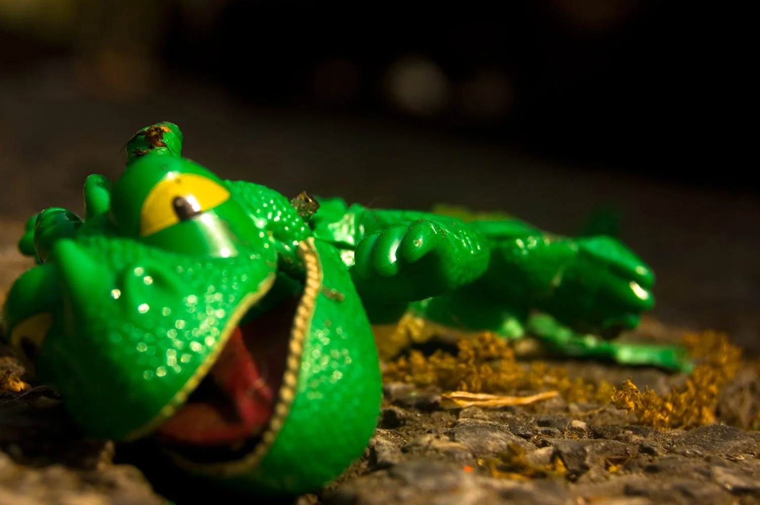 Close-up of a green toy crocodile with an open mouth, lying on a rough surface, with small insects on its head and body, and a dark background.