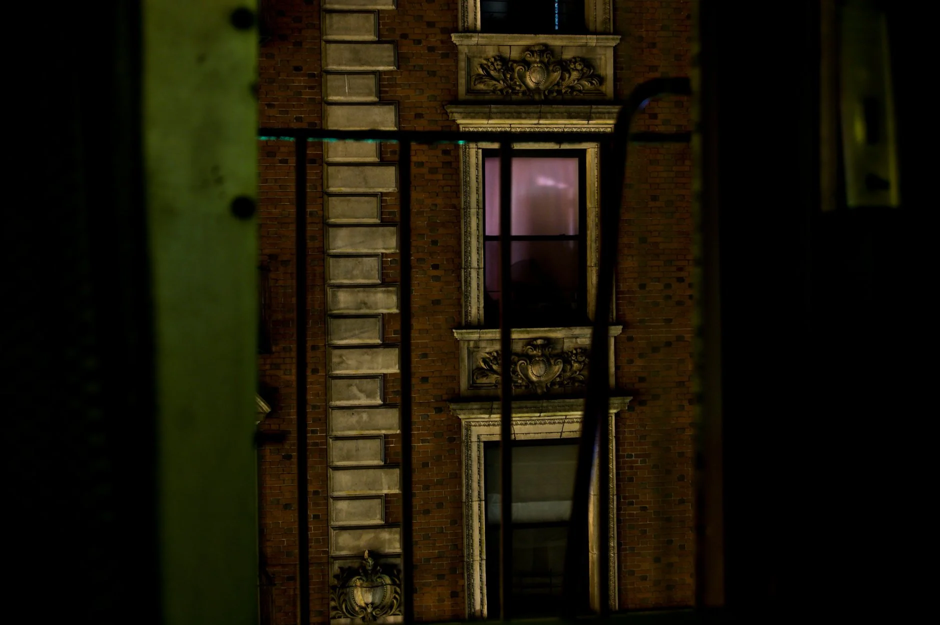 Nighttime view through black metal bars of ornate architectural details on a red brick building, including decorative stone reliefs and window treatments, with a pinkish window reflecting light.