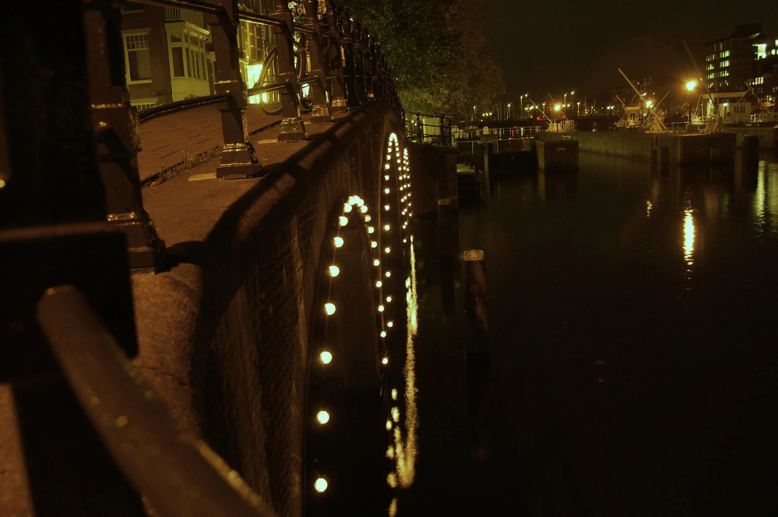 Nighttime view of a dock with a railing decorated with string lights, reflecting on the water. In the background, there are buildings and boats illuminated by streetlights.