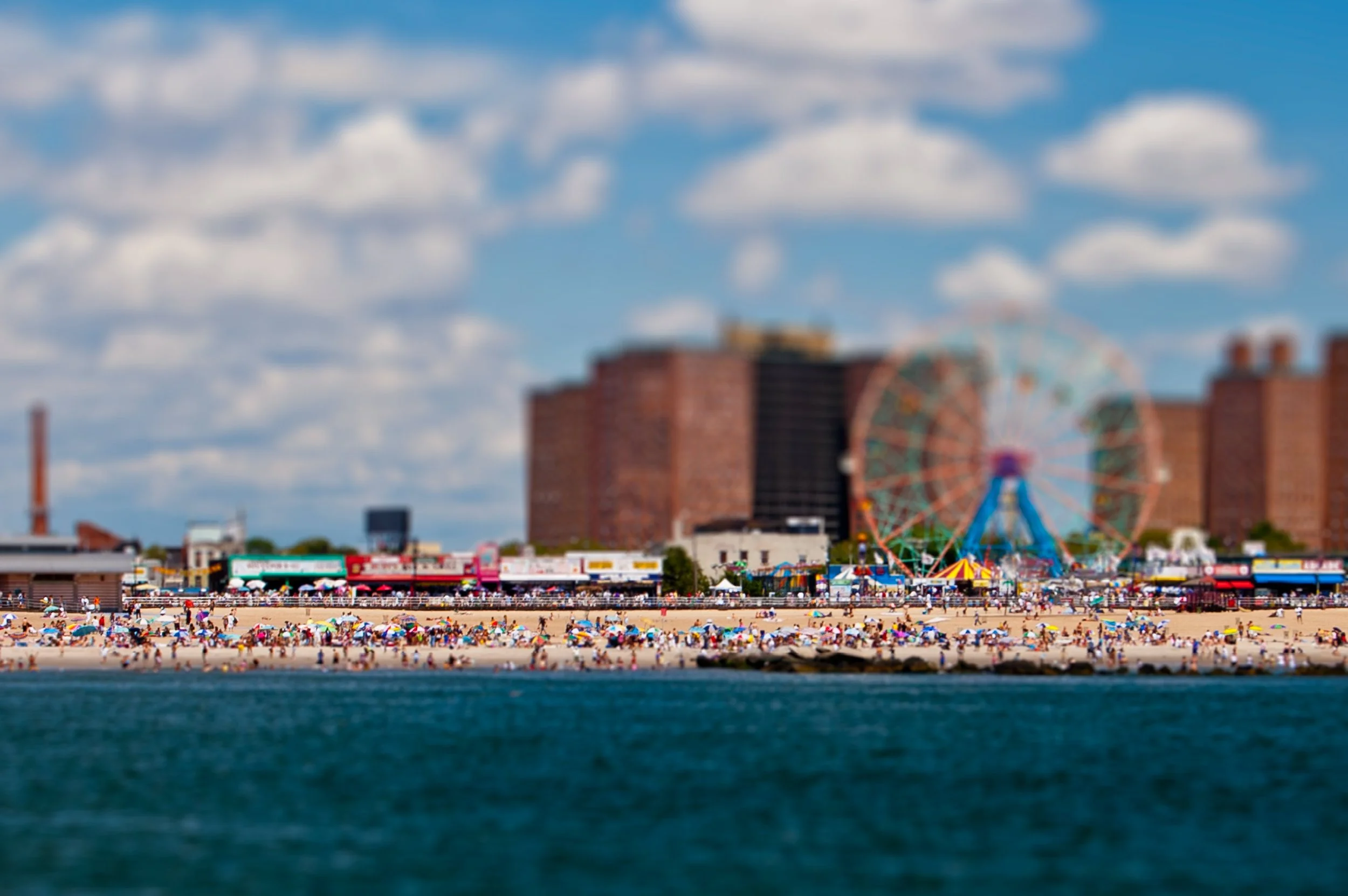 A crowded beach with many people, umbrellas, and beach chairs. In the background, there's a fairground with a ferris wheel and other amusement rides, set against a cityscape with brick buildings and a partly cloudy sky.