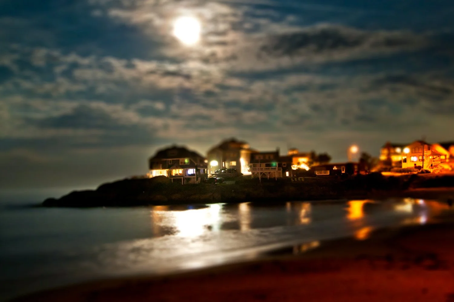 Nighttime scene of houses on a shoreline, illuminated by warm lights, with a full moon in a cloudy sky reflected on the water.