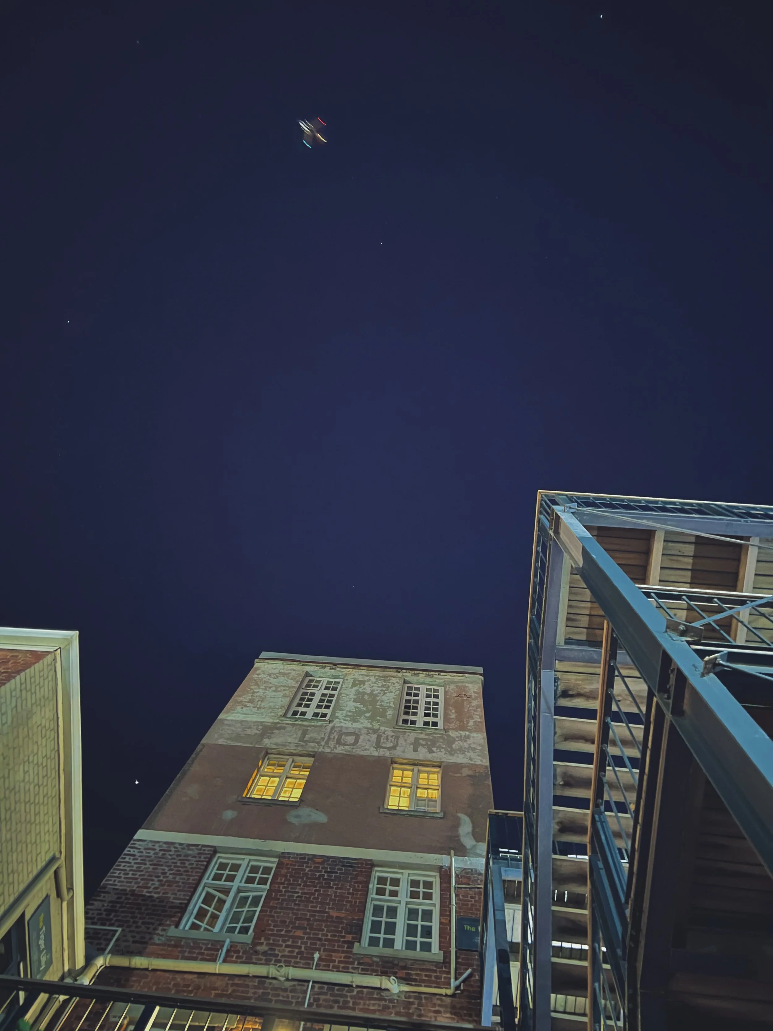Night view looking up at the sky with a building's facade and balconies below. The building has illuminated windows and brick exterior, and there is a metal staircase on the right side of the image.