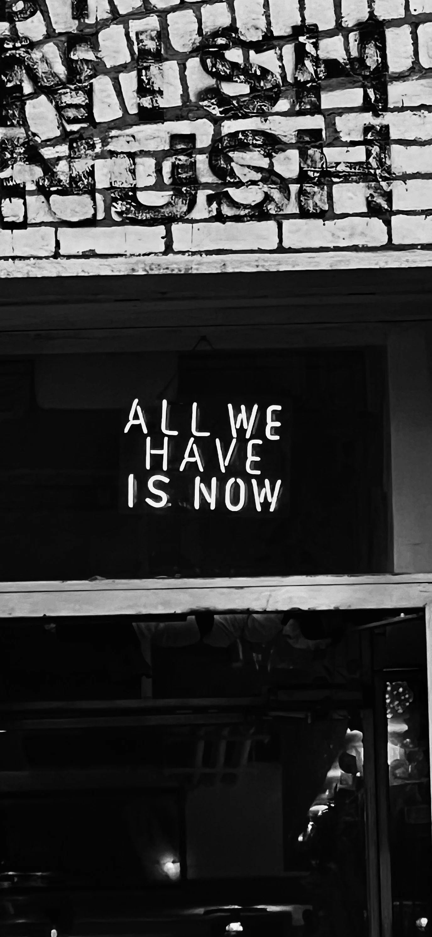 Black and white photo of a brick wall with graffiti and a neon sign that reads 'ALL WE HAVE IS NOW'.