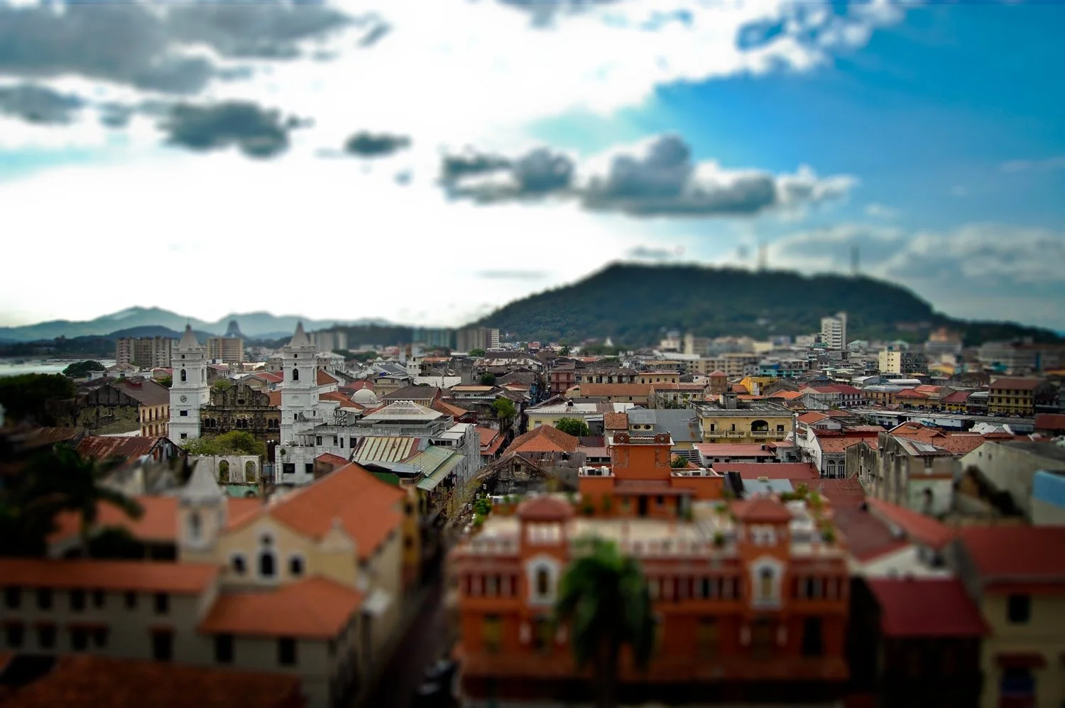 A cityscape with a mix of colonial and modern buildings, houses with red-tiled roofs, and a prominent church with twin bell towers in the foreground. A hill with wind turbines is in the background under a partly cloudy sky.