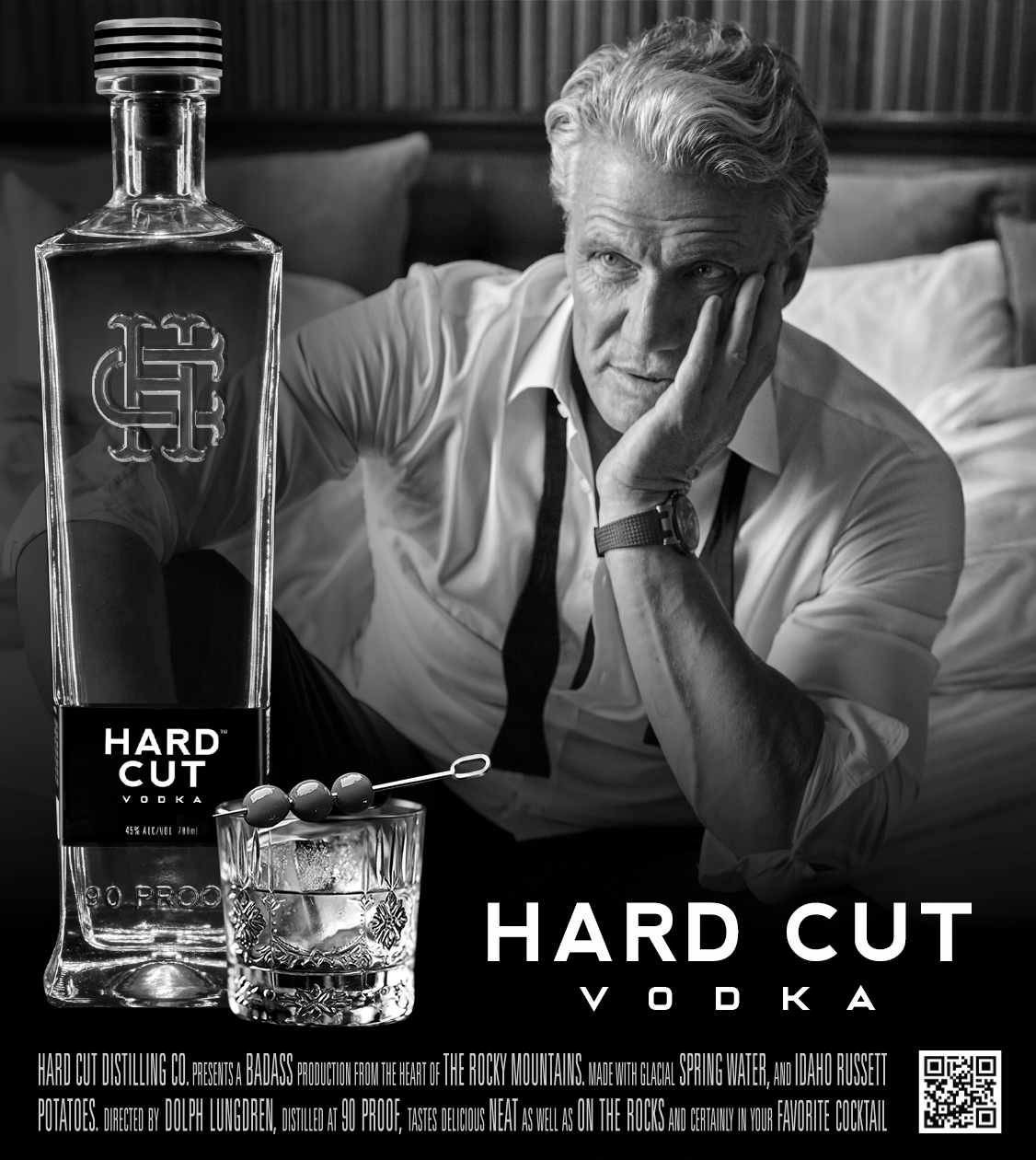 Black and white photo of a man with platinum blonde hair, wearing a white shirt, sitting on a bed with a contemplative expression, holding his face with one hand. Next to him is a large bottle of Hard Cut Vodka, a glass with ice, and a cocktail pick with olives.