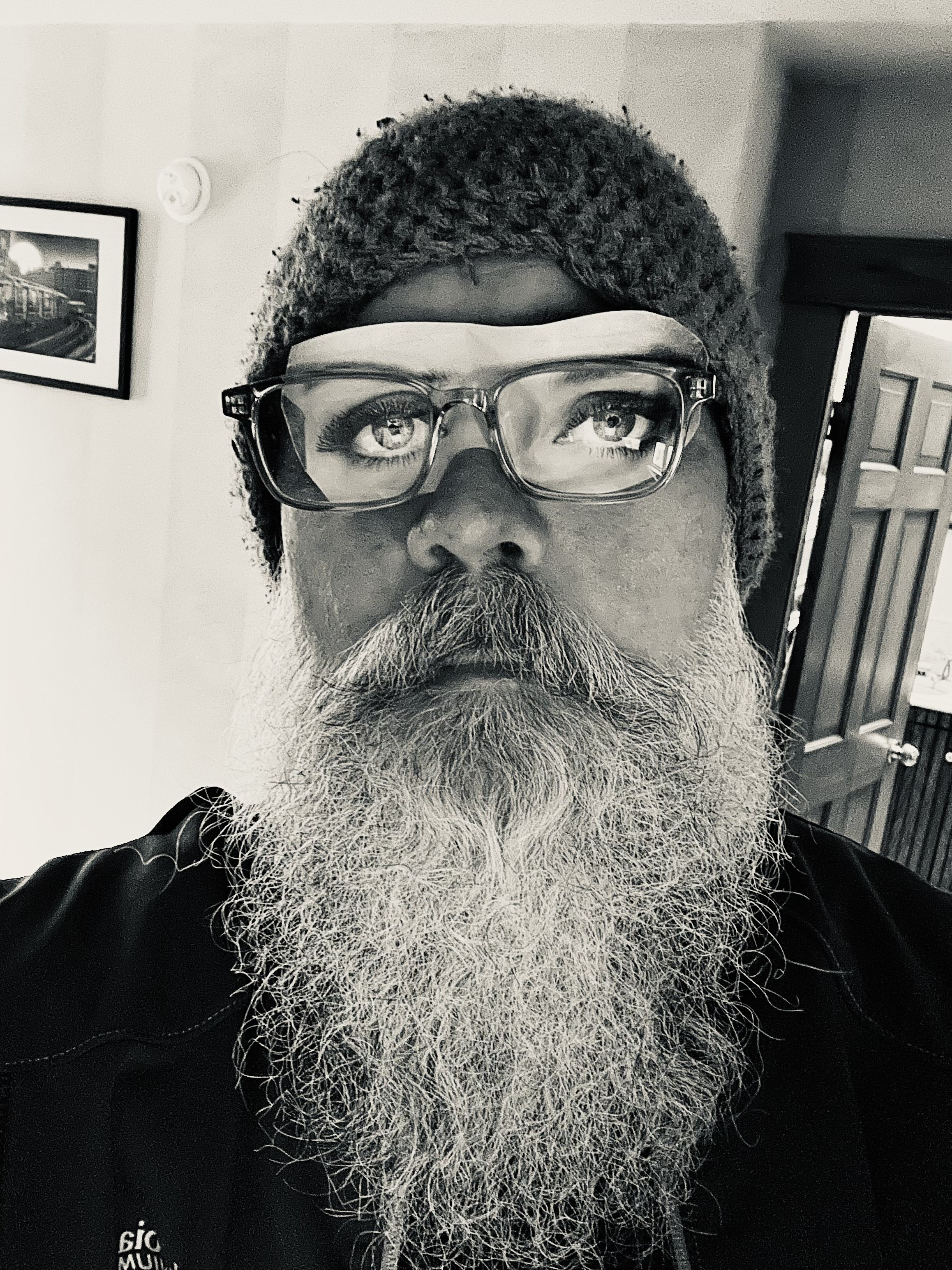 A close-up black and white photo of a man with a long, curly beard wearing clear eyeglasses and a knitted hat, standing indoors near a door and a framed picture on the wall.