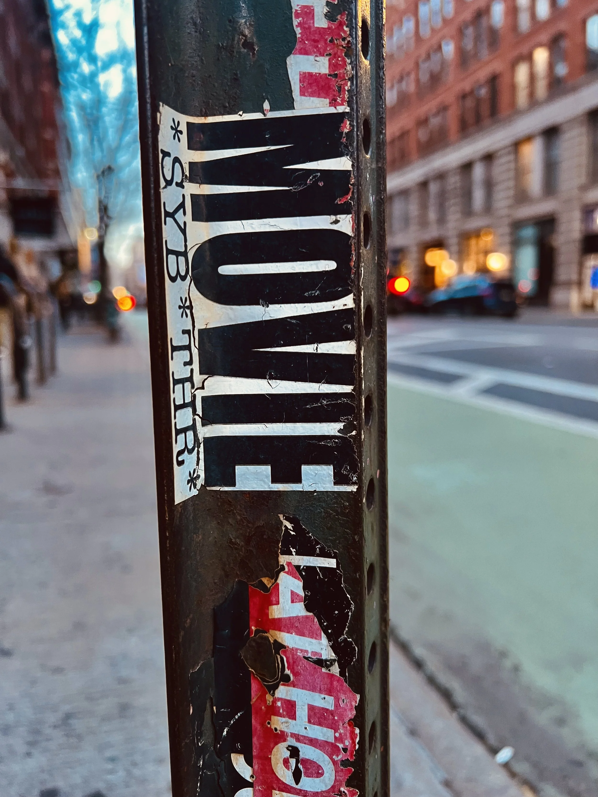 Close-up of a worn, peeling sticker on a black metal post with a city street in the background featuring buildings, cars, and blurred lights.