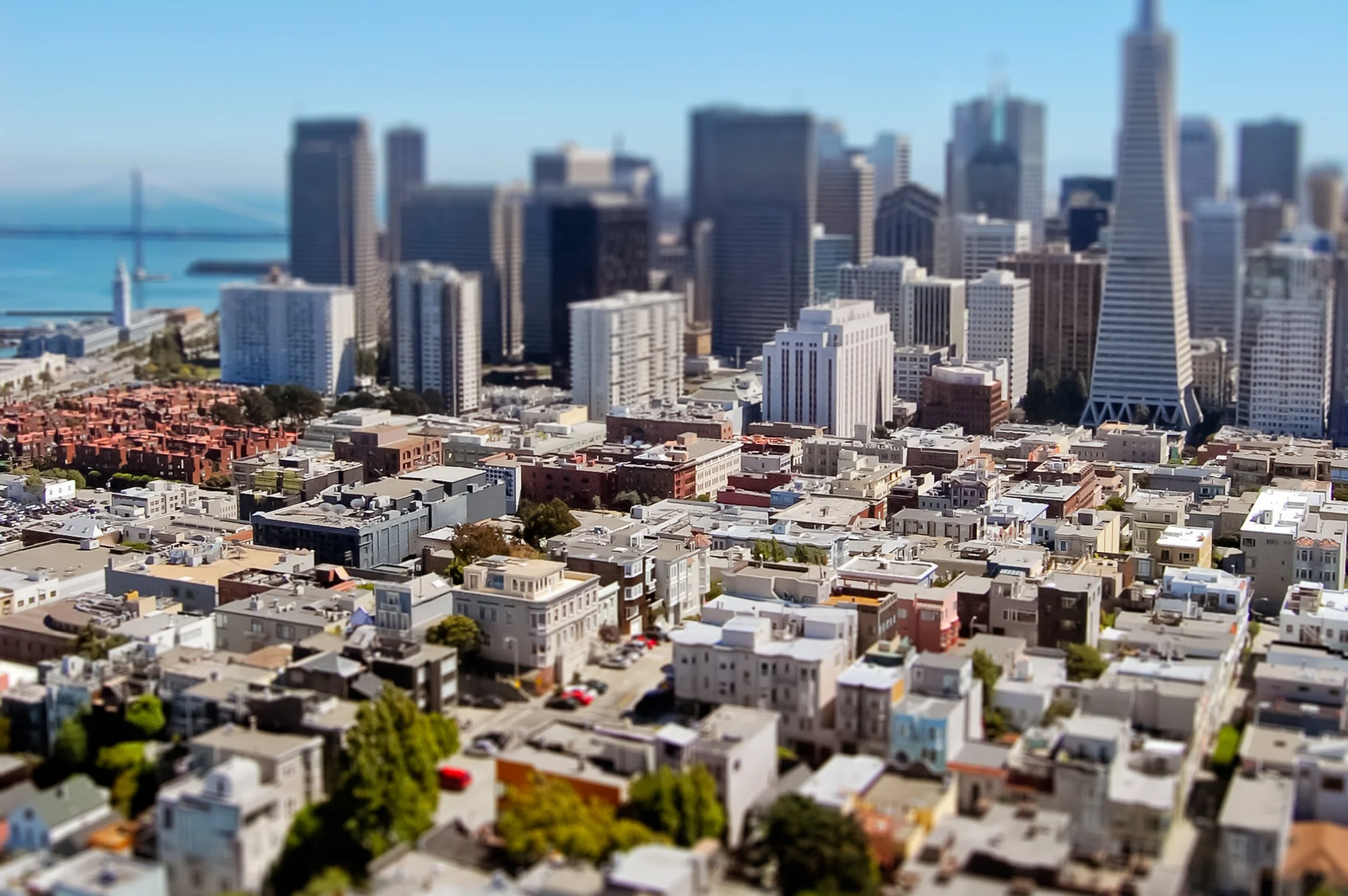 Aerial cityscape view of San Francisco skyline with skyscrapers, buildings, and a bridge over water in the background, with a tilt-shift effect.