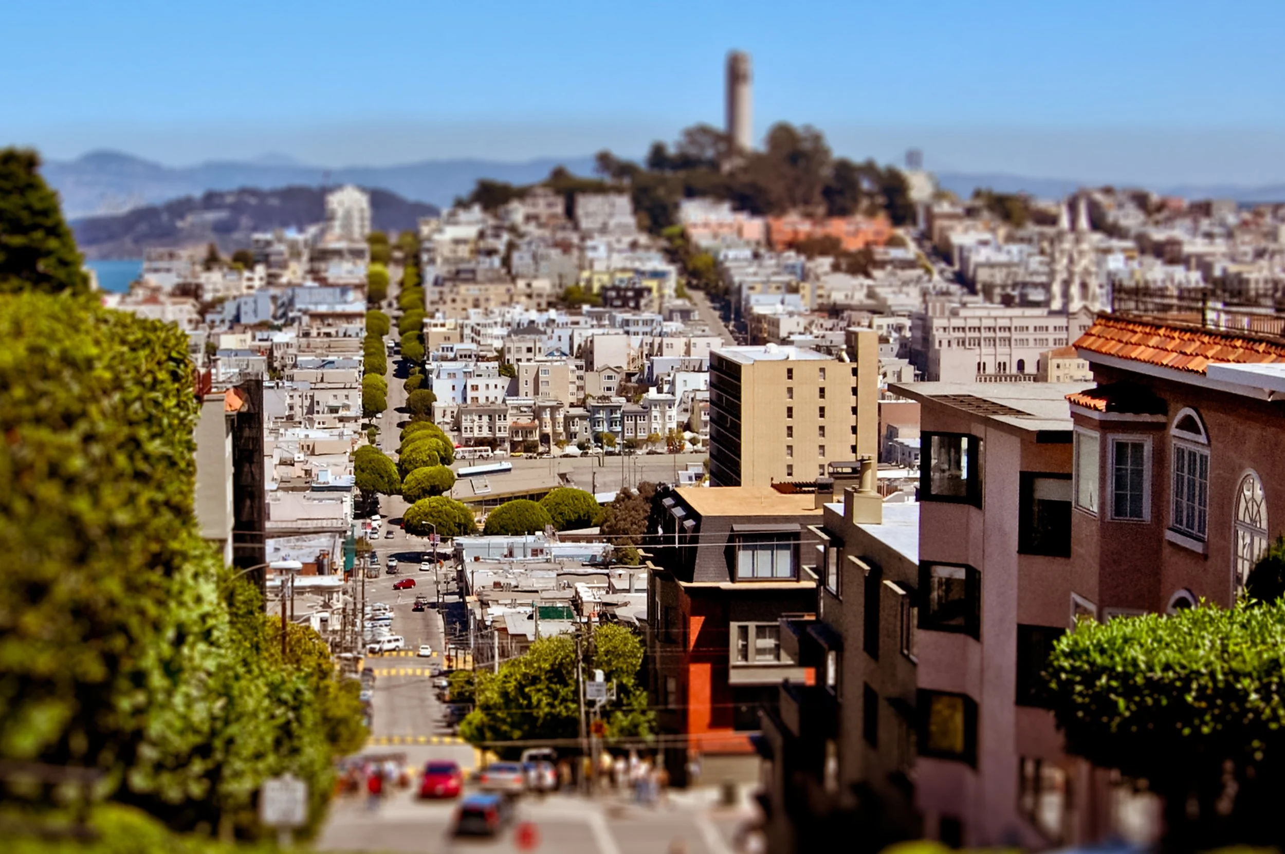 San Francisco cityscape featuring a hill with a tall tower at the top, residential buildings, and greenery in the foreground under a clear blue sky.