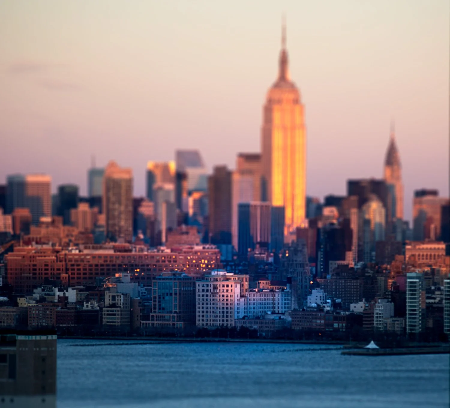 Sunset view of the New York City skyline with the Empire State Building illuminated and reflected in the water.