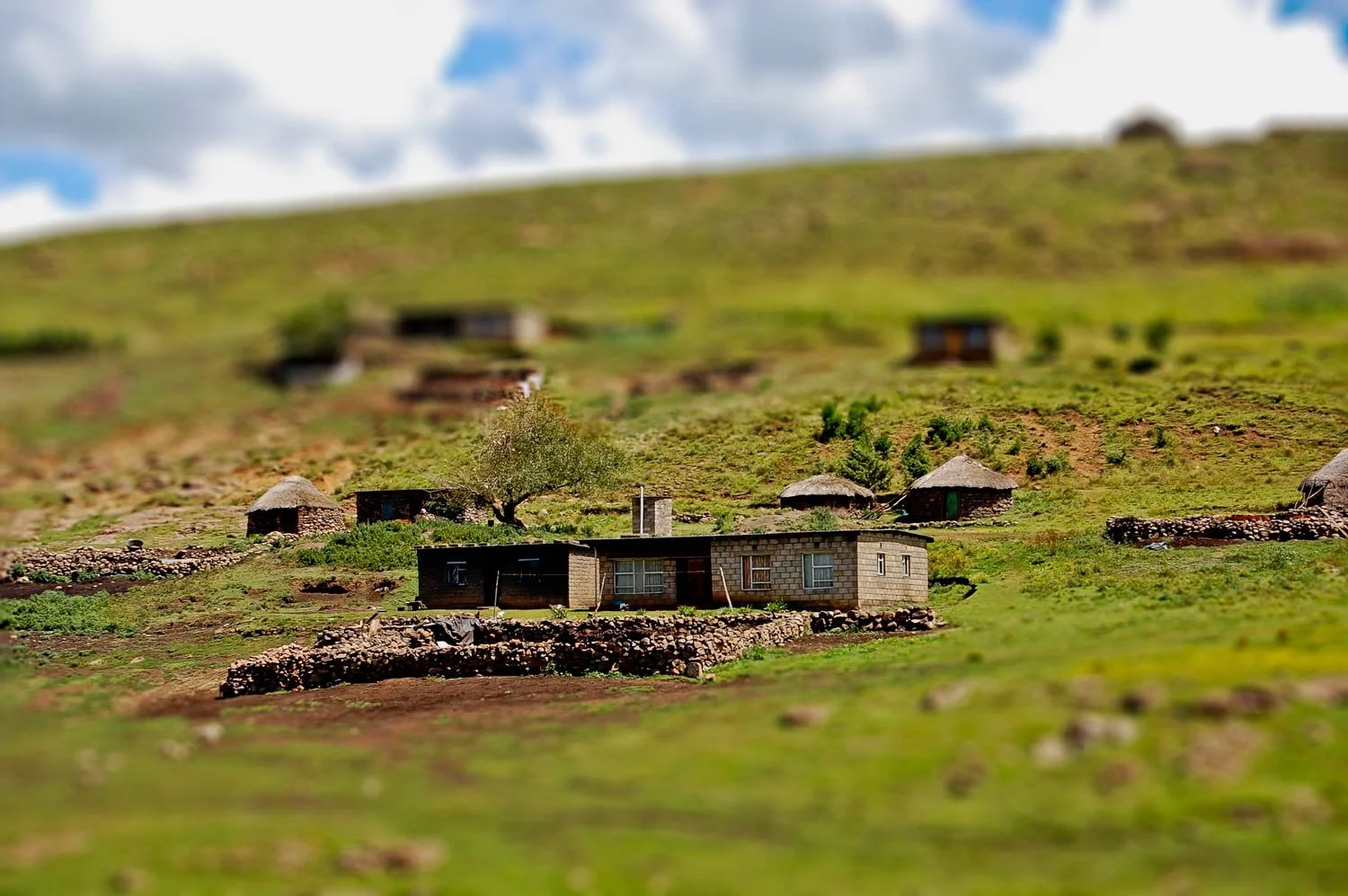 A small rural village with traditional huts, a modern brick house, a tree, and stone fences on a green hillside under a cloudy sky.