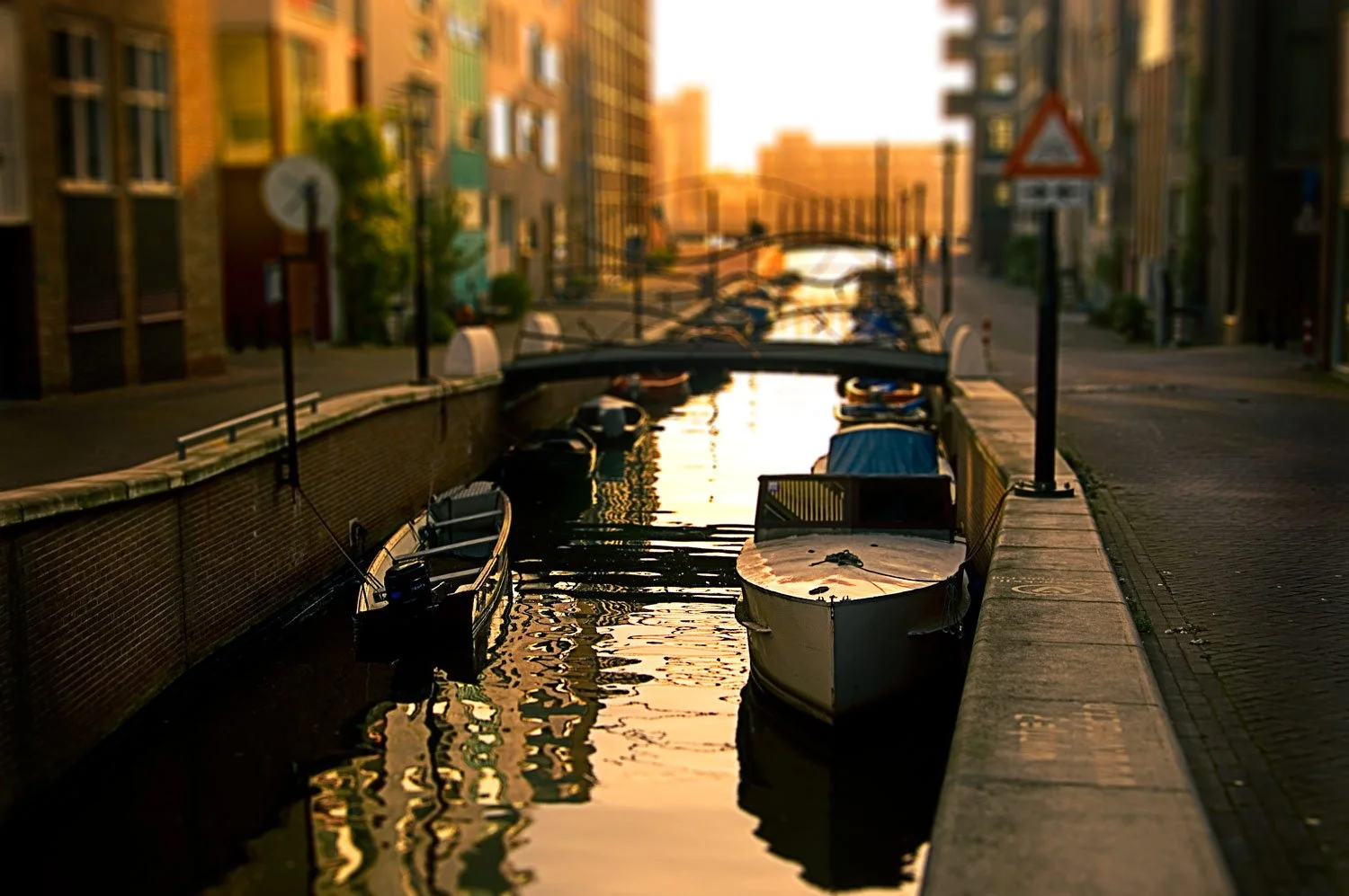 Boats docked along a canal with a bridge in the background during sunset, in an urban area with buildings on either side.