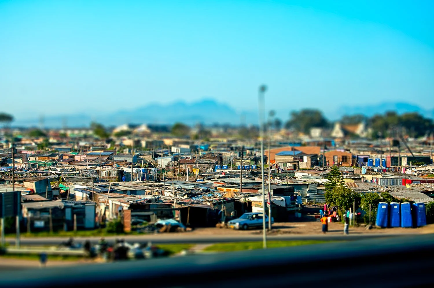 A densely populated area with makeshift houses and shanties, some people visible on the street, portable toilets on the right side, and mountains in the background, under a clear blue sky.