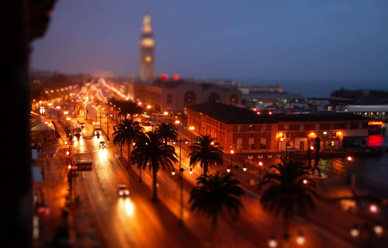 A nighttime city street scene with palm trees, streetlights, moving vehicles, a building with lit windows, and a blurred iconic clock tower in the background.