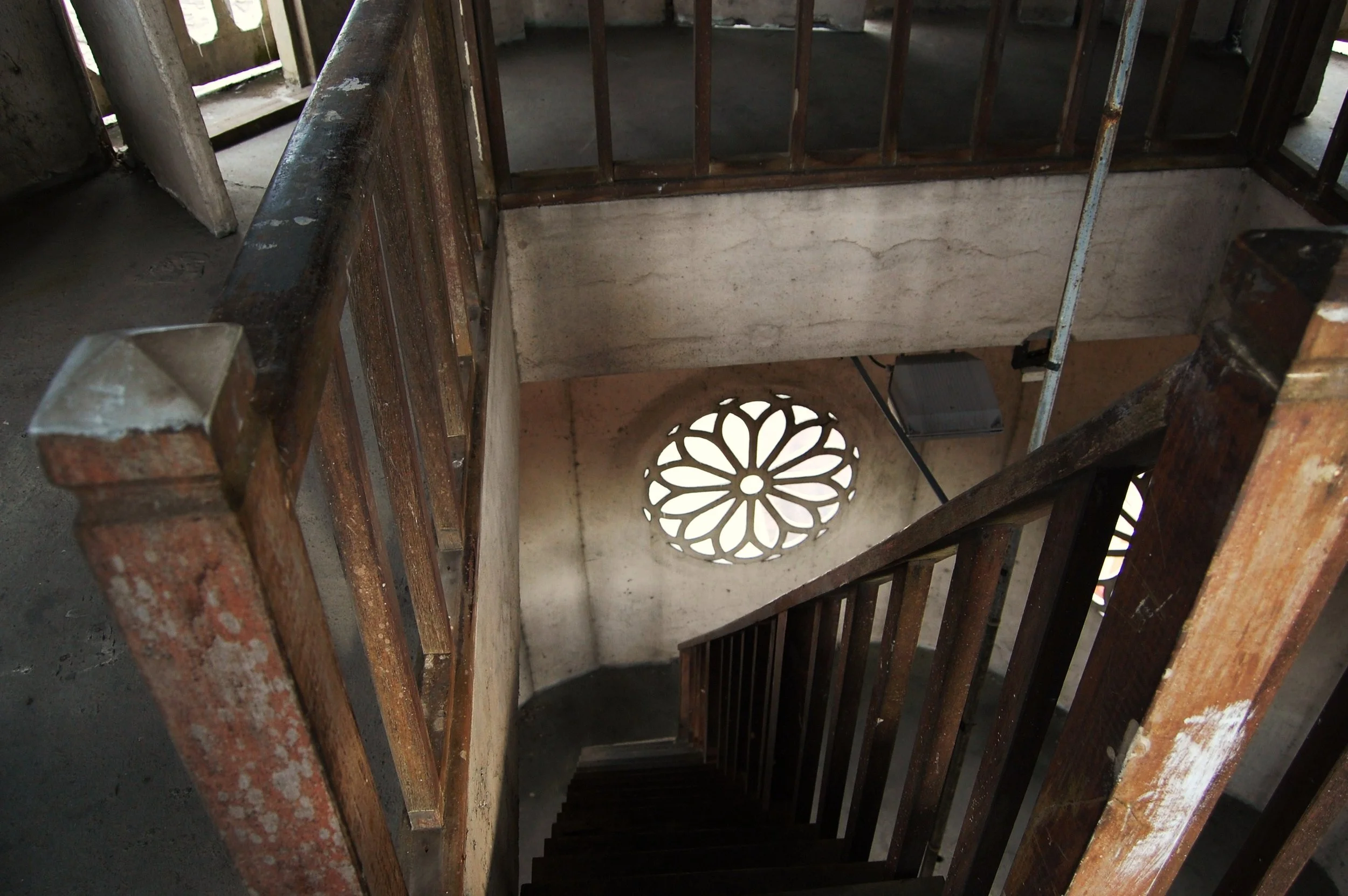 Rusty wooden staircase with a decorative circular window at the bottom and an outdoor opening at the top allowing natural light to enter.